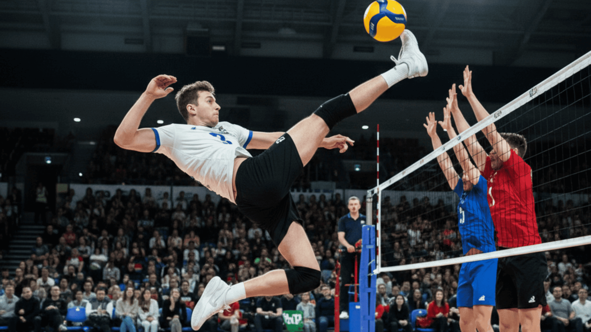 white jersey volleyball player jumps and kicks the ball over the net as two blockers jump in a packed arena