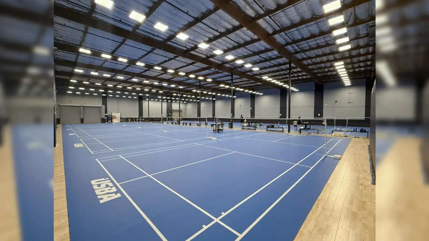 wide view of empty indoor badminton courts with blue flooring and bright overhead lights in a large sports facility