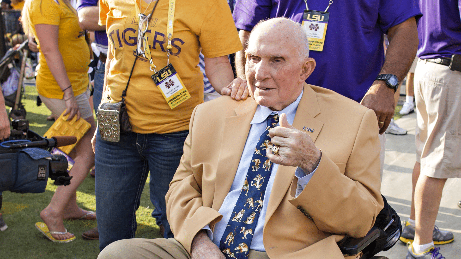 _y.a. tittle in a tan suit sits in a wheelchair giving a thumbs up as LSU fans stand around him outdoors
