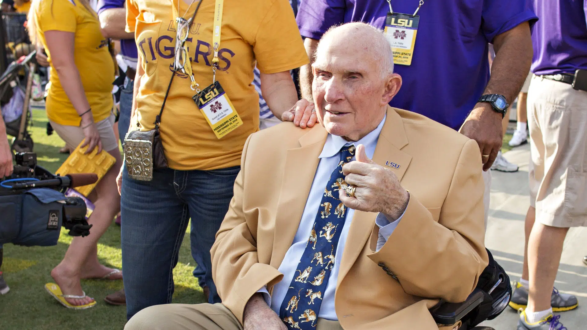 _y.a. tittle in a tan suit sits in a wheelchair giving a thumbs up as LSU fans stand around him outdoors