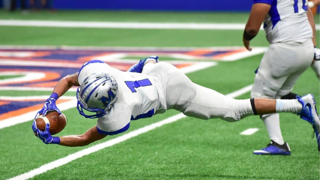 A football player in a white uniform dives forward to catch the ball near the end zone on a green field turff