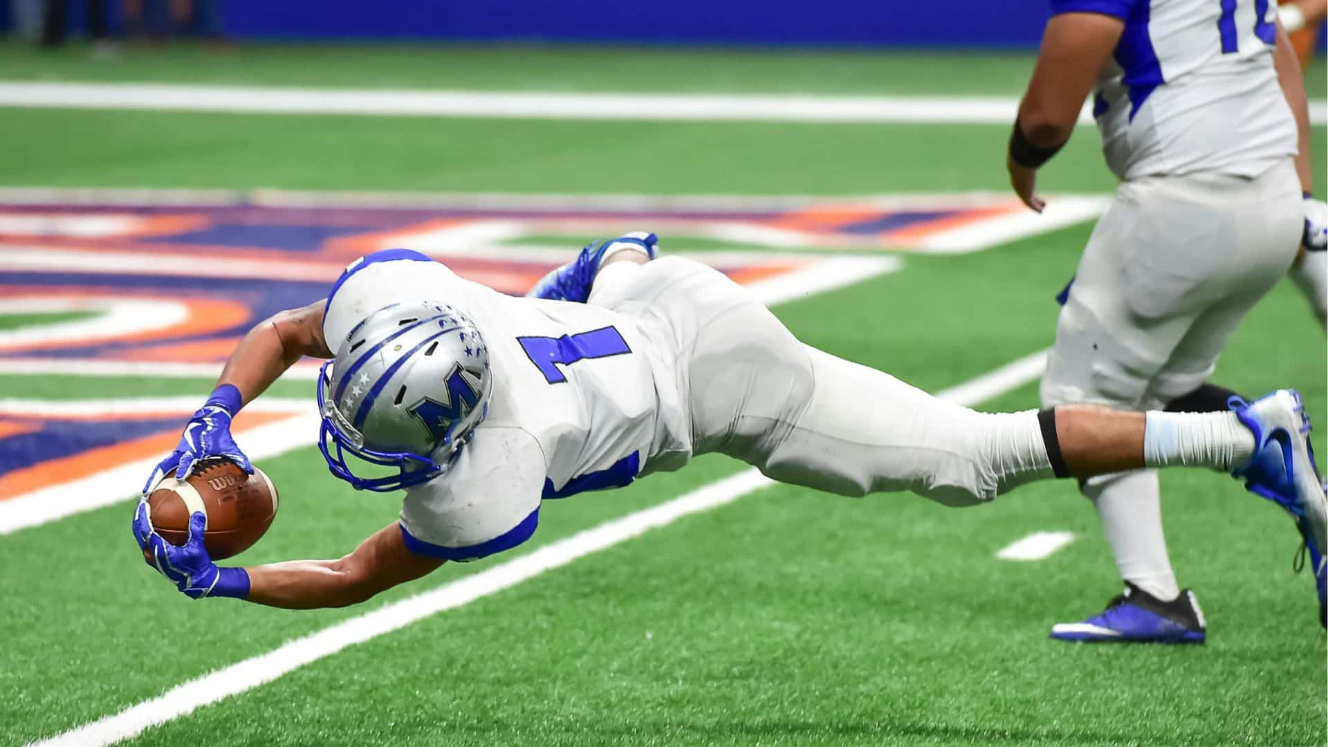 A football player in a white uniform dives forward to catch the ball near the end zone on a green field turff