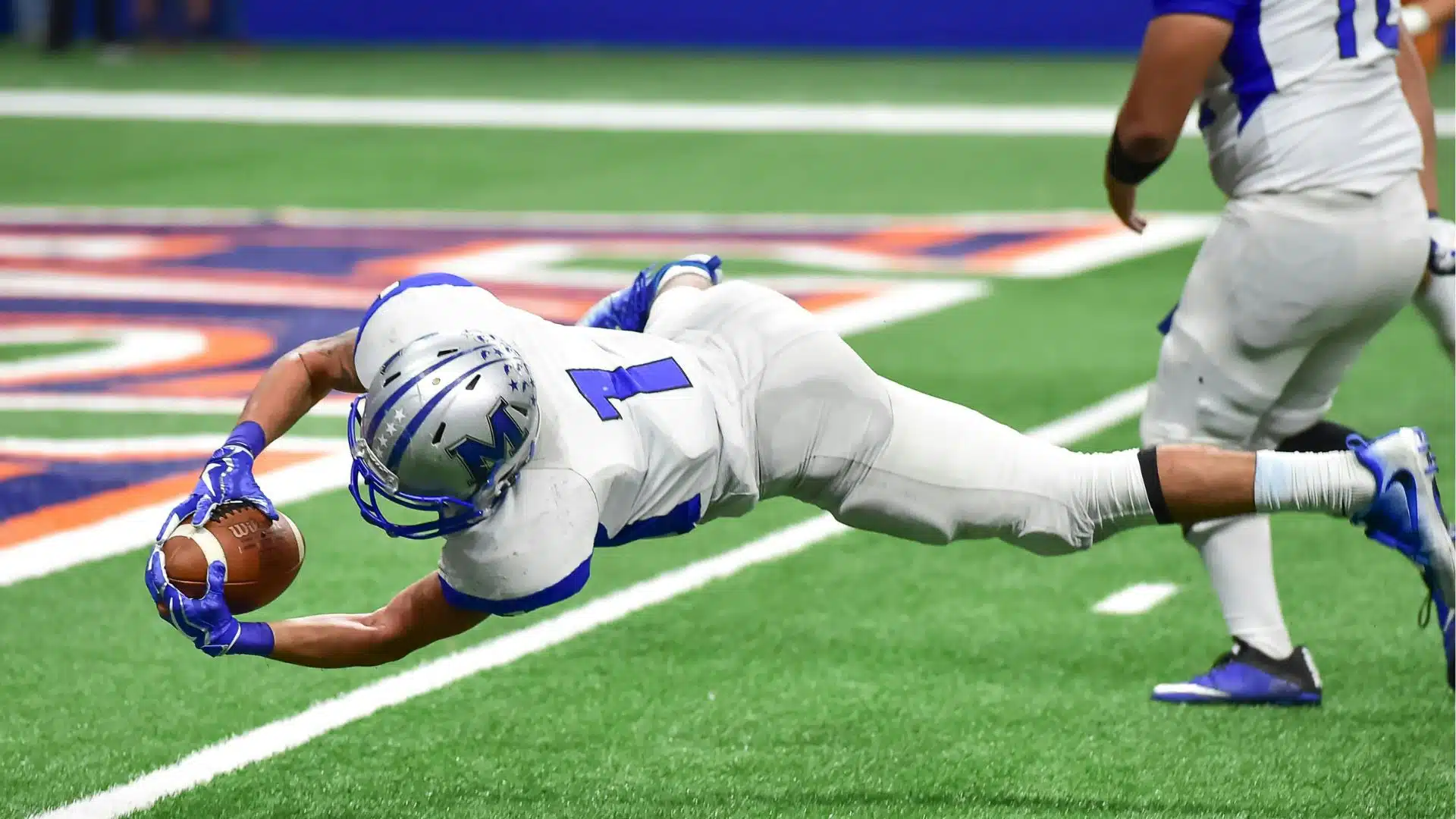 A football player in a white uniform dives forward to catch the ball near the end zone on a green field turff