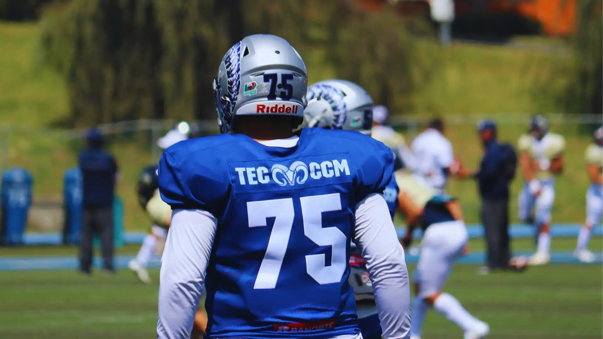 A football player wearing a blue jersey with number 75 stands on the field during practice as teammates run drills behind him