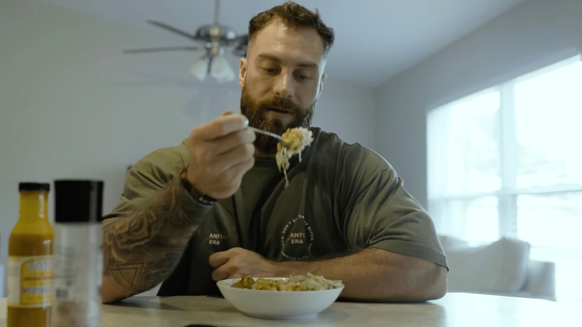 a bearded man sitting at a table eating from a bowl with a fork in a bright kitchen with a window behind him