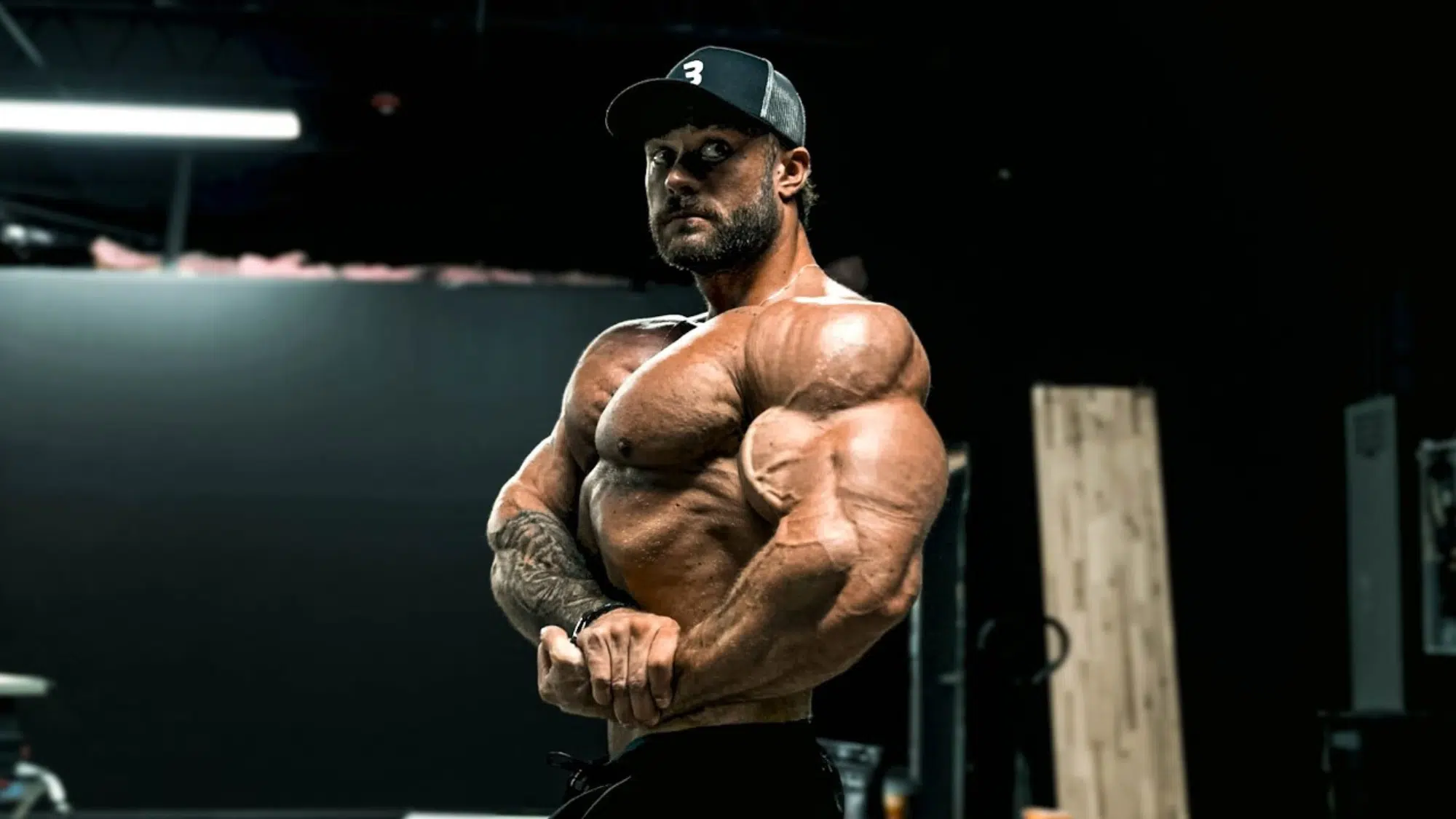 a muscular man wearing a black cap posing and flexing his arms in a dimly lit gym