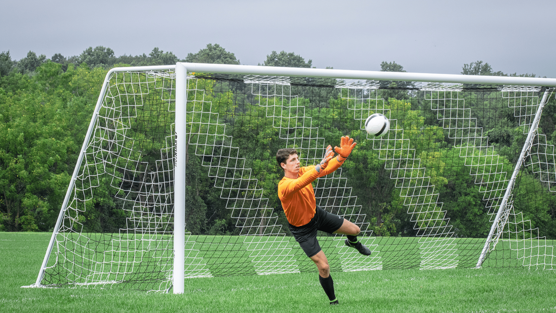 a soccer goalkeeper in orange jersey diving to block a ball in front of the goal net on a grassy field