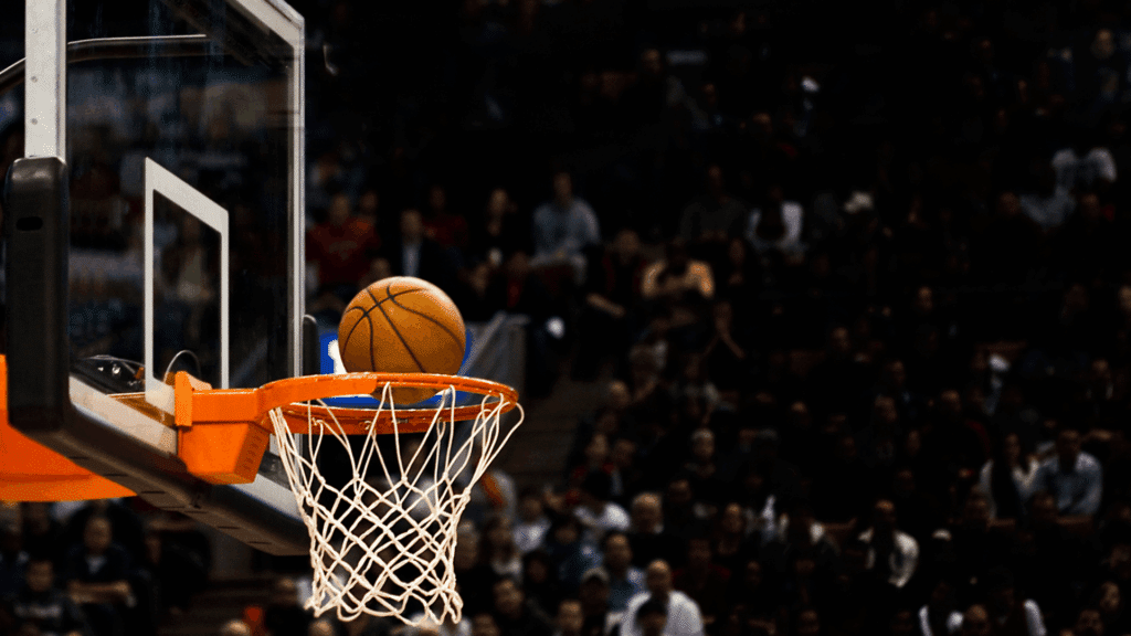 basketball going in the rim during a game with hoop and net in focus and a blurred crowd in the arena background