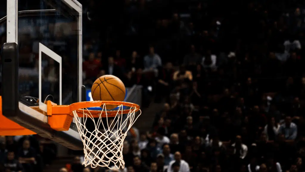 basketball going in the rim during a game with hoop and net in focus and a blurred crowd in the arena background