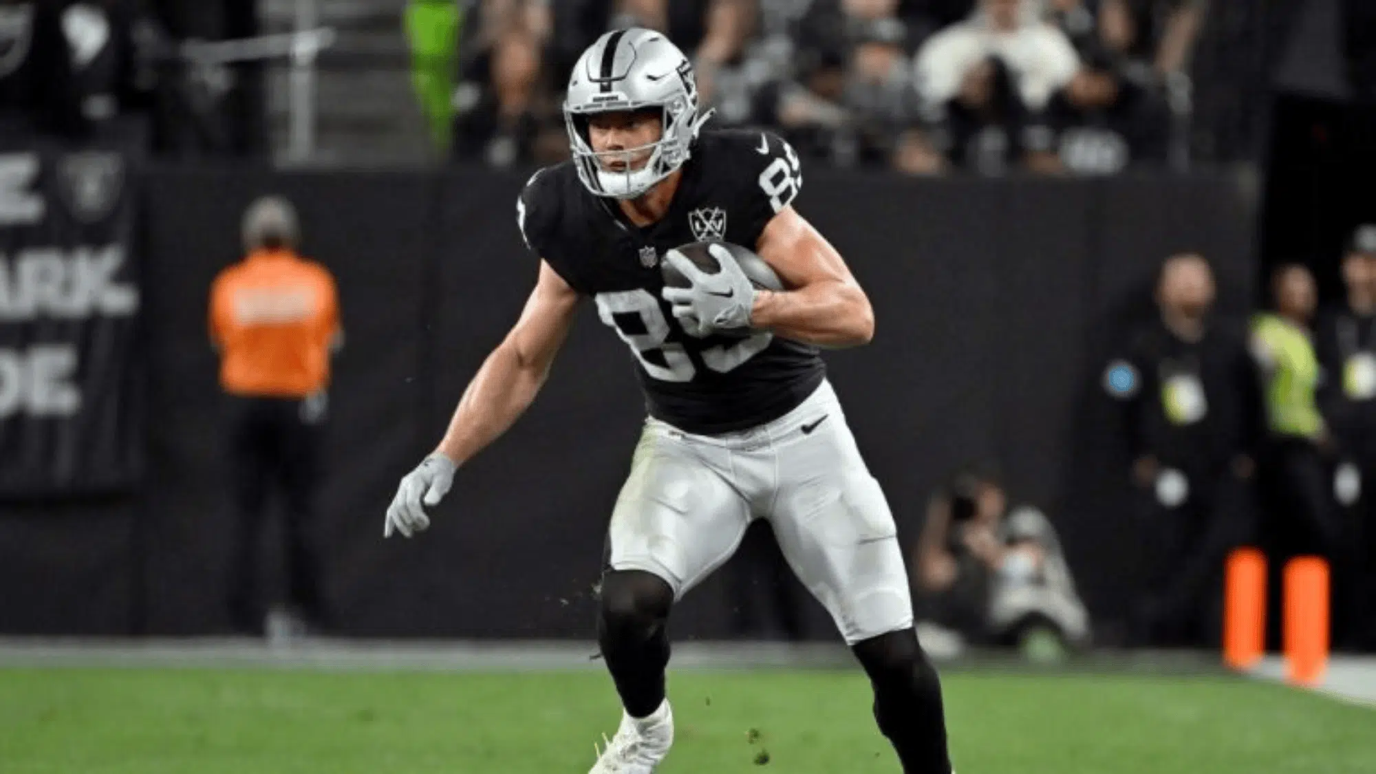 brock bowers in a black and silver uniform runs on a field, holding a football