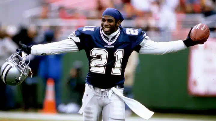 deion sanders in a blue jersey with number 21 smiles jubilantly, holding a helmet in one hand and a football in the other on a field