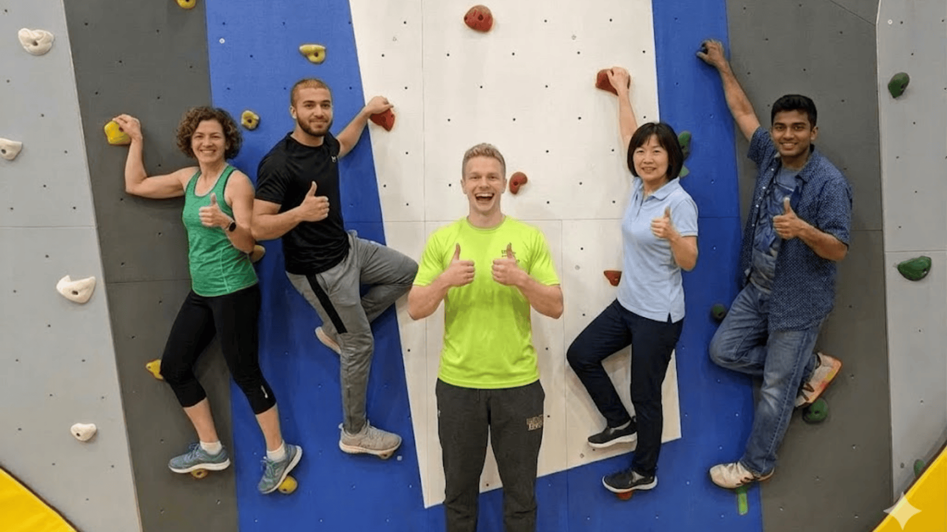 five people smiling and climbing an indoor rock wall while a man in front gives thumbs up to camera