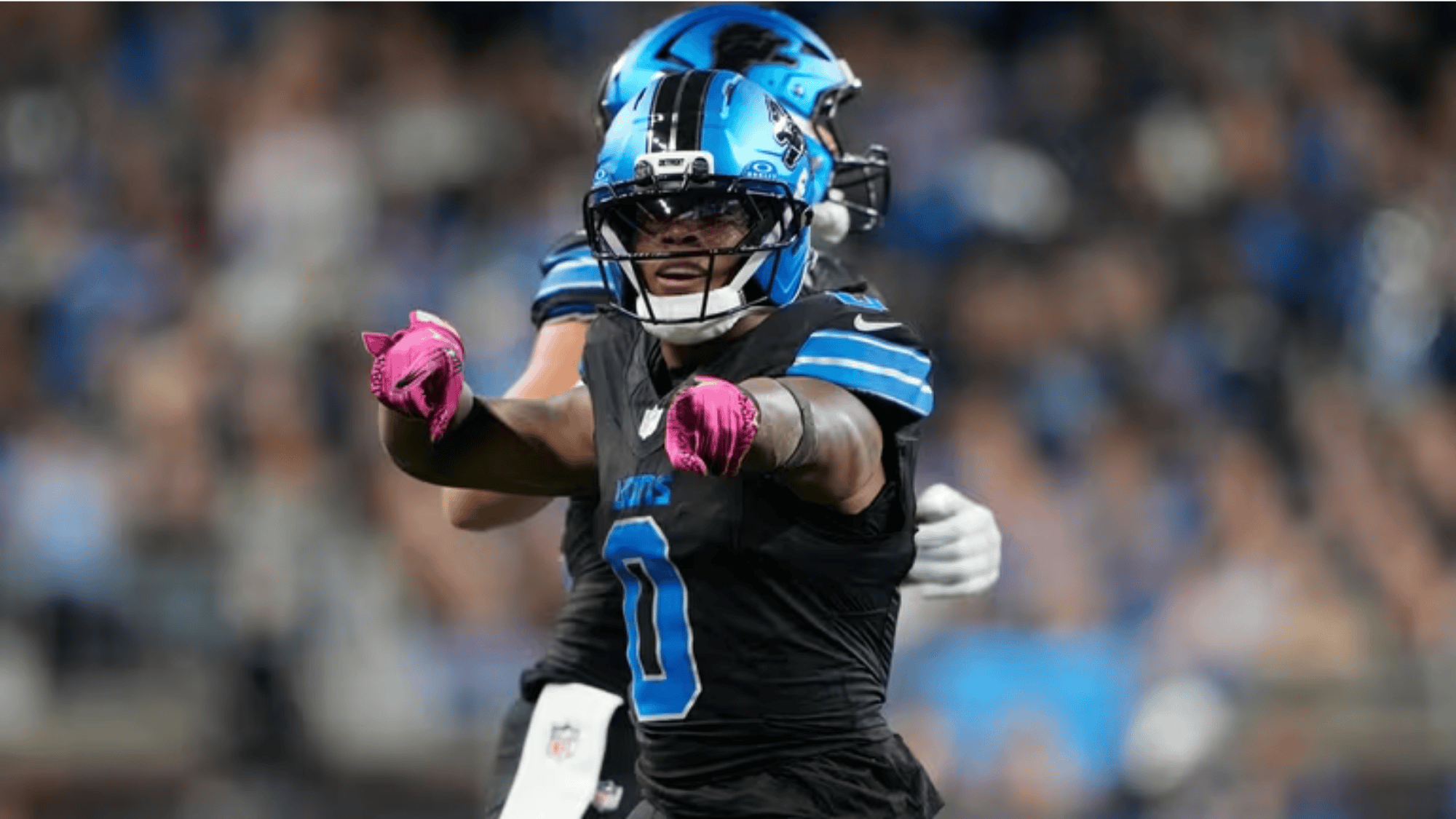 jahmyr gibbs in a black and blue uniform and helmet gestures energetically with pink gloves on