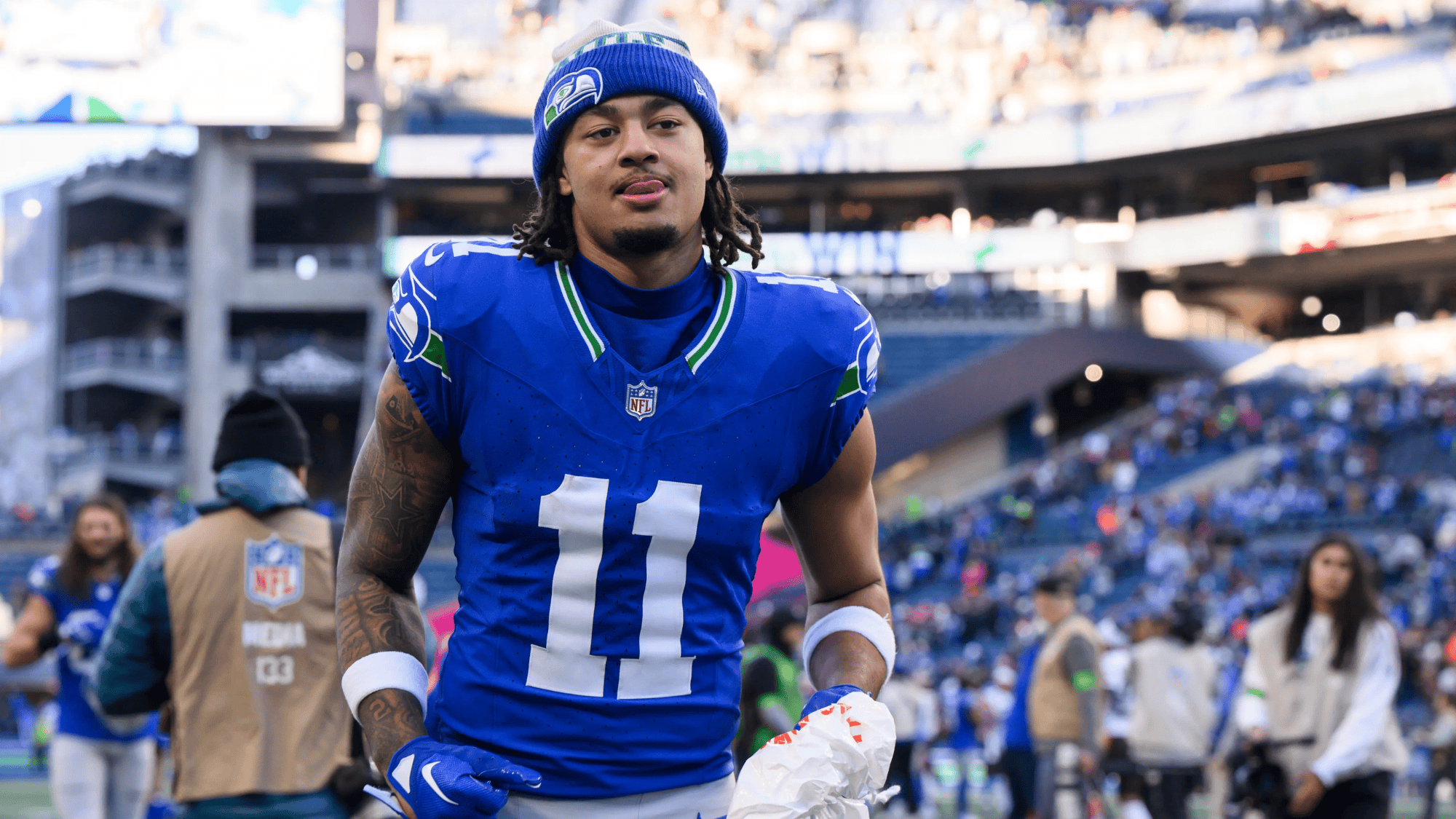 jaxon smith-njigba in a blue jersey and beanie walks on a stadium field holding white items