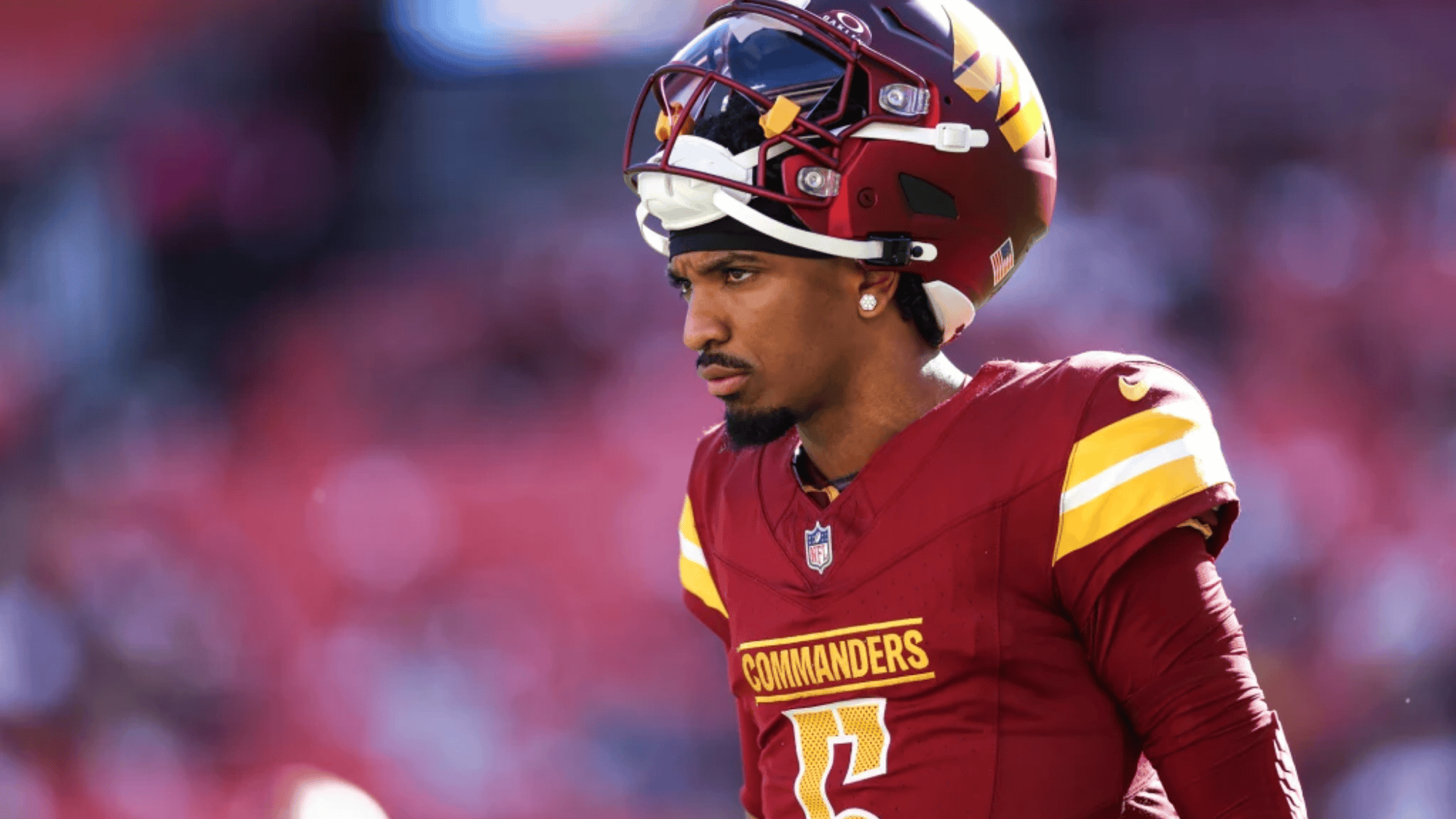 jayden daniels in a red commanders uniform with yellow accents stands on a blurred field looking focused