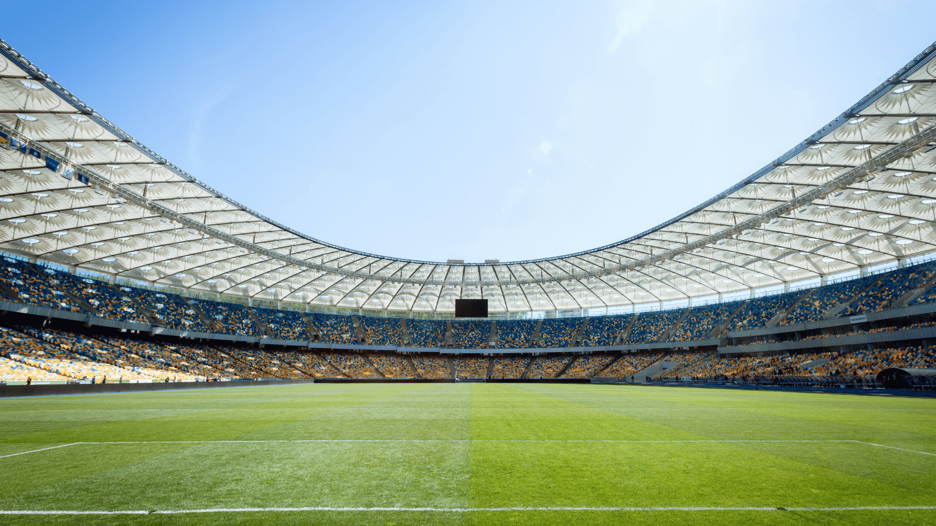 large open stadium with curved roof rows of seats and green field under bright sky