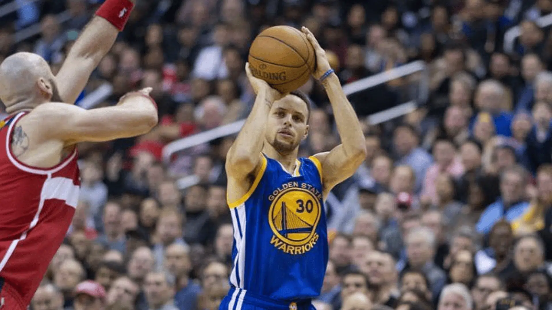 stephen curry in a warriors jersey shooting a three pointer during an nba game with a defender closing in and crowd blurred behind