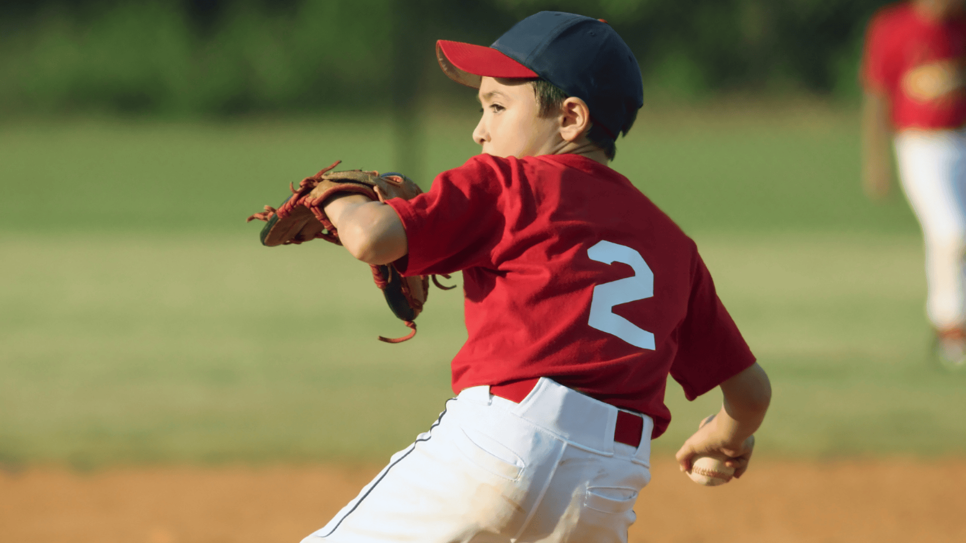 young boy in red baseball uniform pitching a ball on a sunny field wearing a glove and cap captured mid throw action