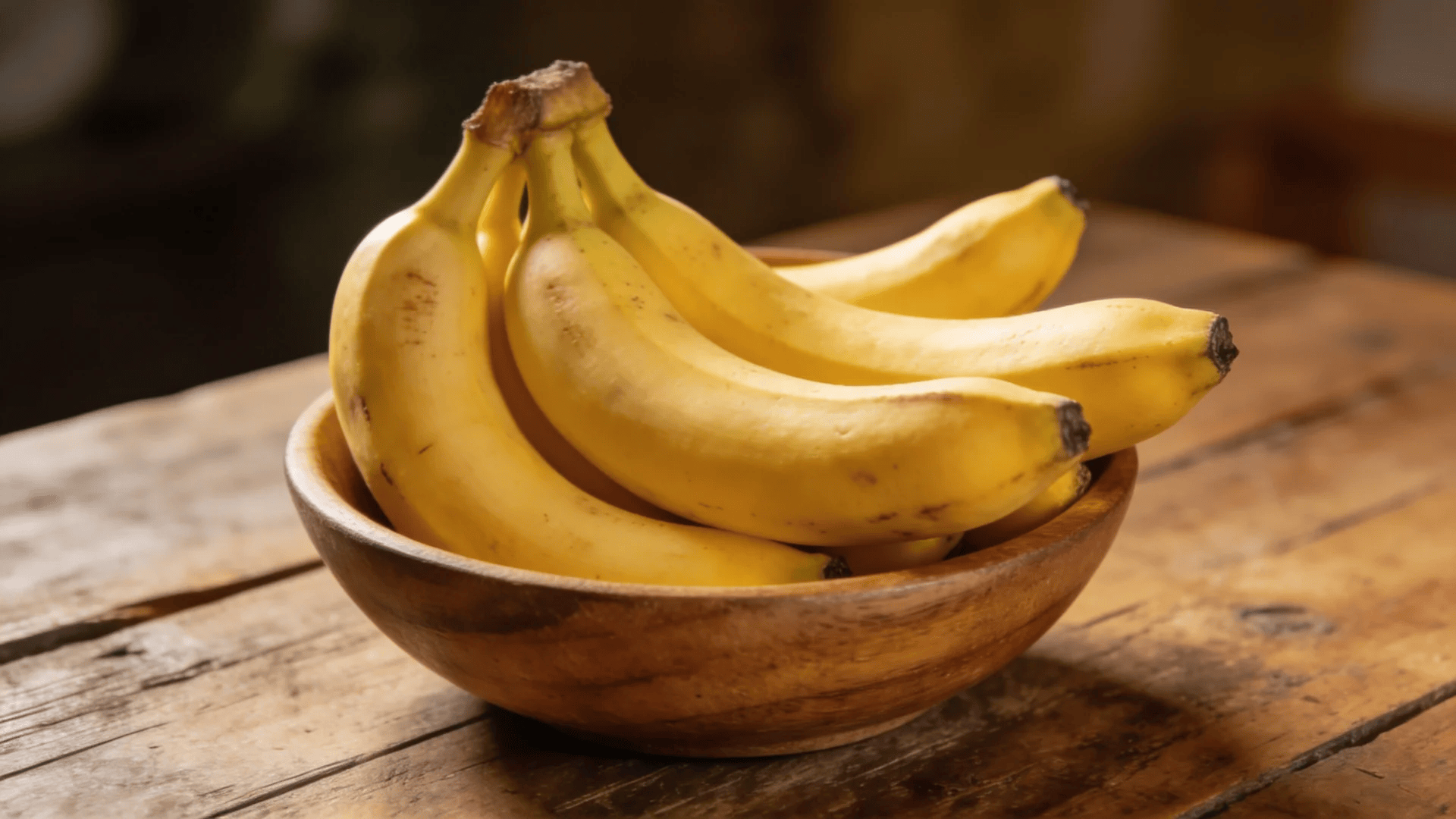 a bunch of ripe yellow bananas resting in a wooden bowl placed on a rustic wooden table in warm indoor lighting