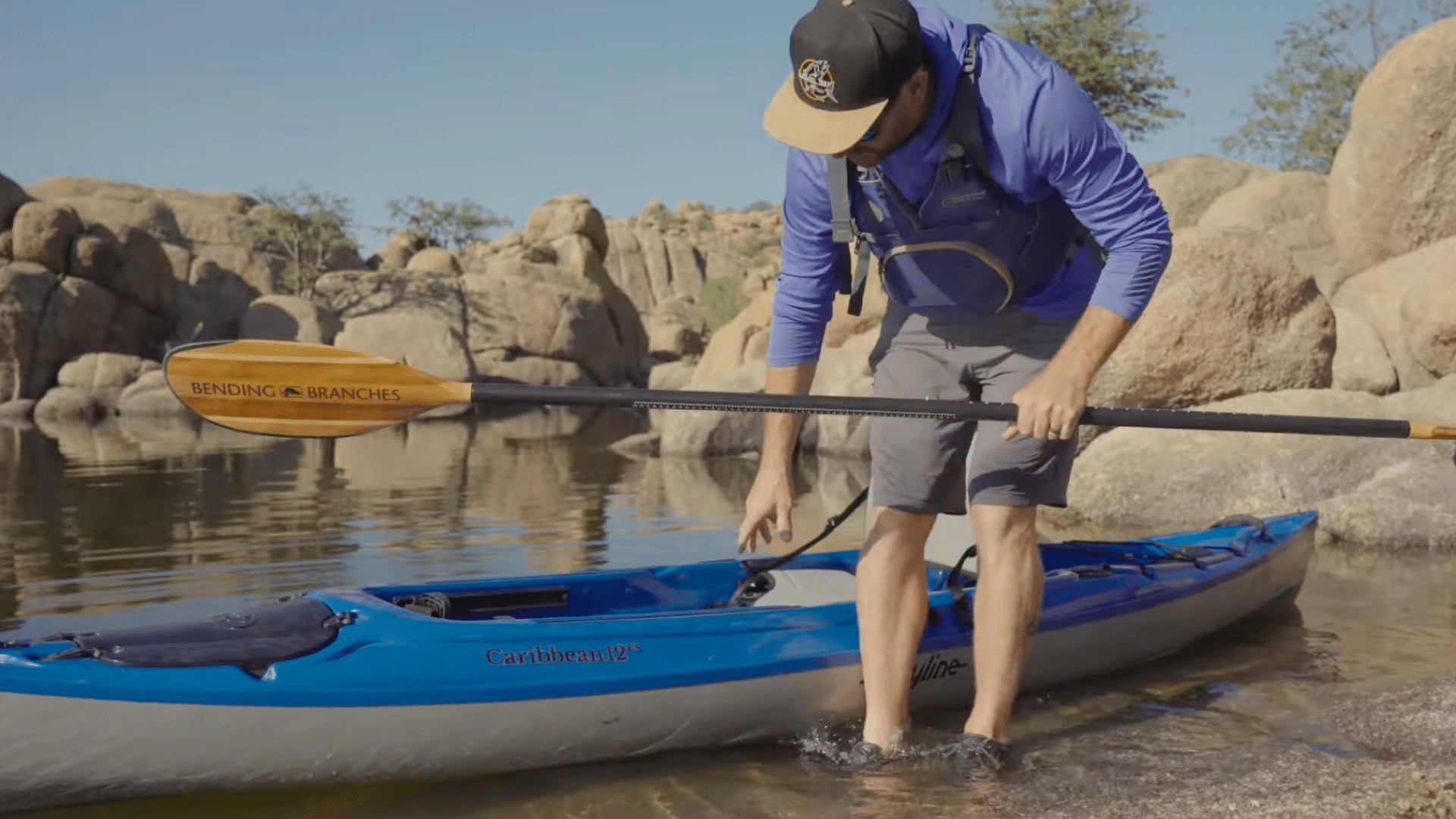 a man stands in shallow water holding a kayak steady while positioning himself carefully to get into the kayak safely