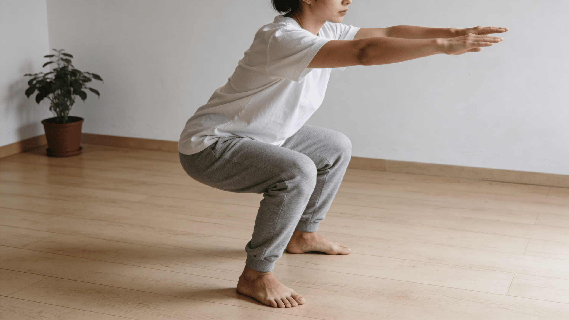 person performing squat exercise indoors with arms extended wearing casual clothes on wooden floor near plant