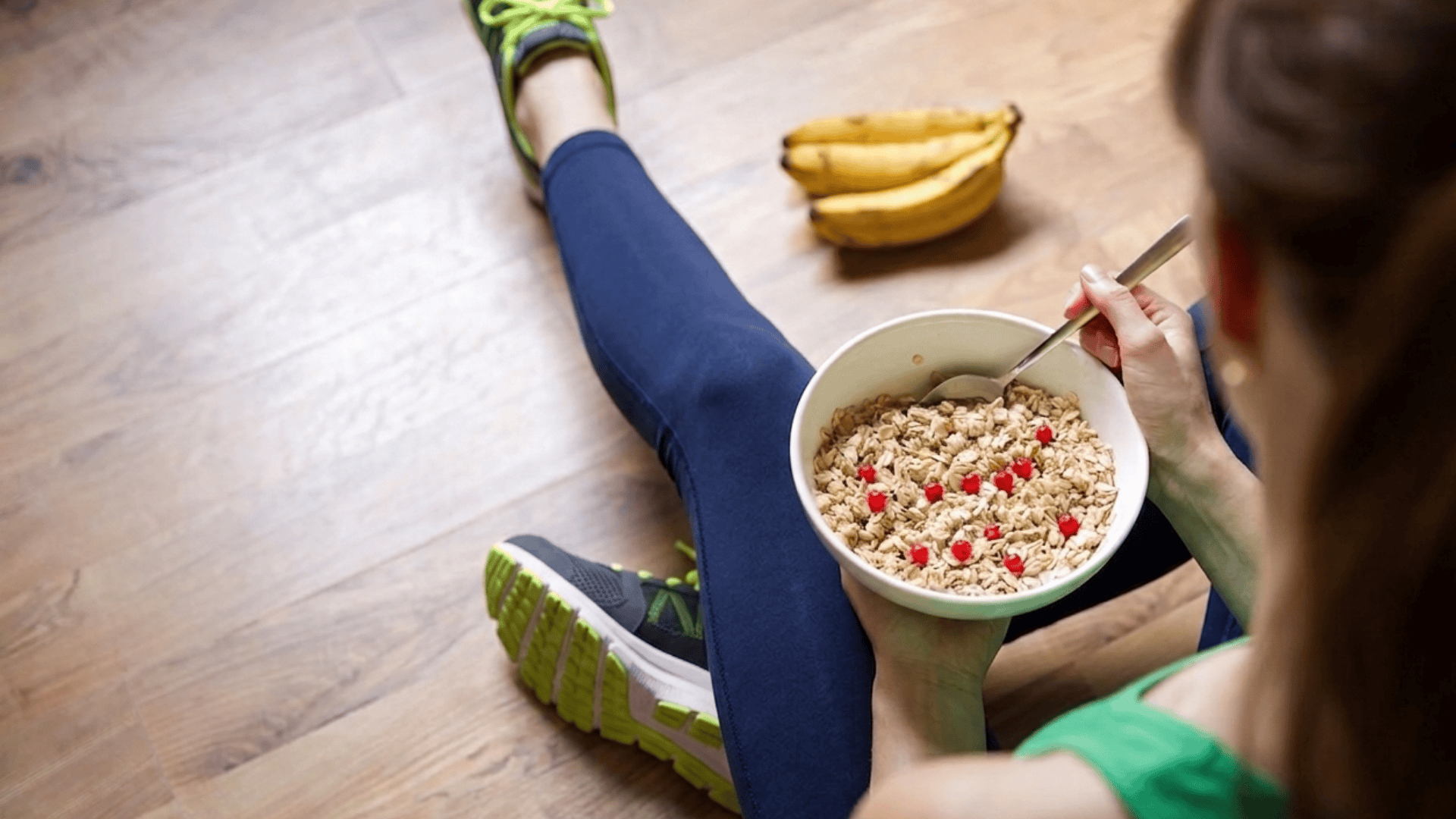 person sitting on floor after workout eating oats with berries with bananas nearby wearing running shoes