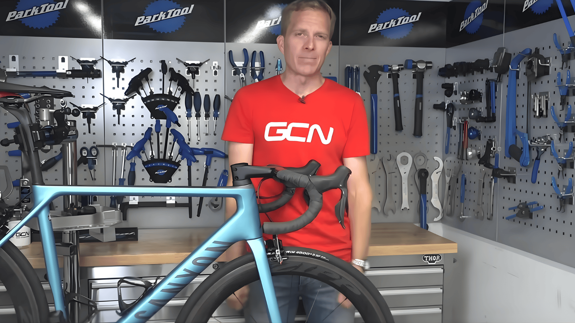 person standing in bike workshop next to road bike explaining how to change bike tire with tools organized on wall behind