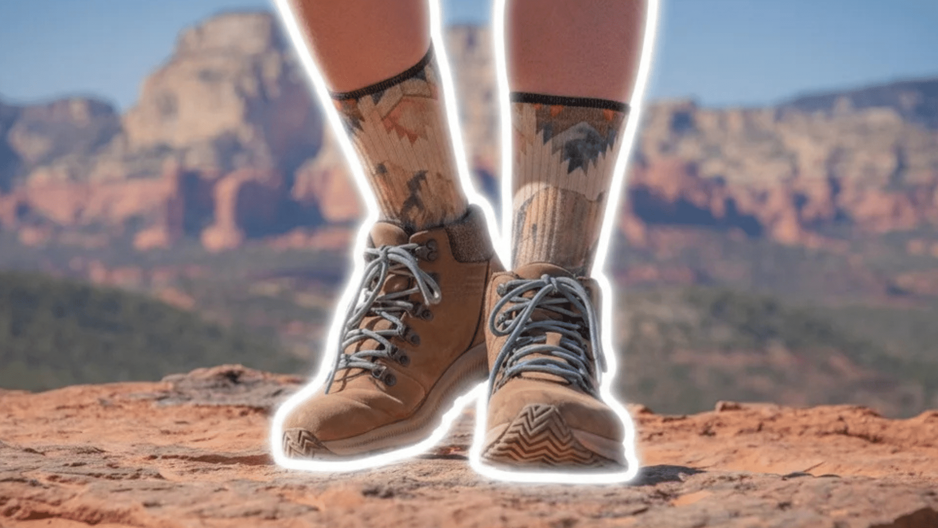 person standing on rocky ledge wearing brown boots and patterned socks with desert canyon landscape in background