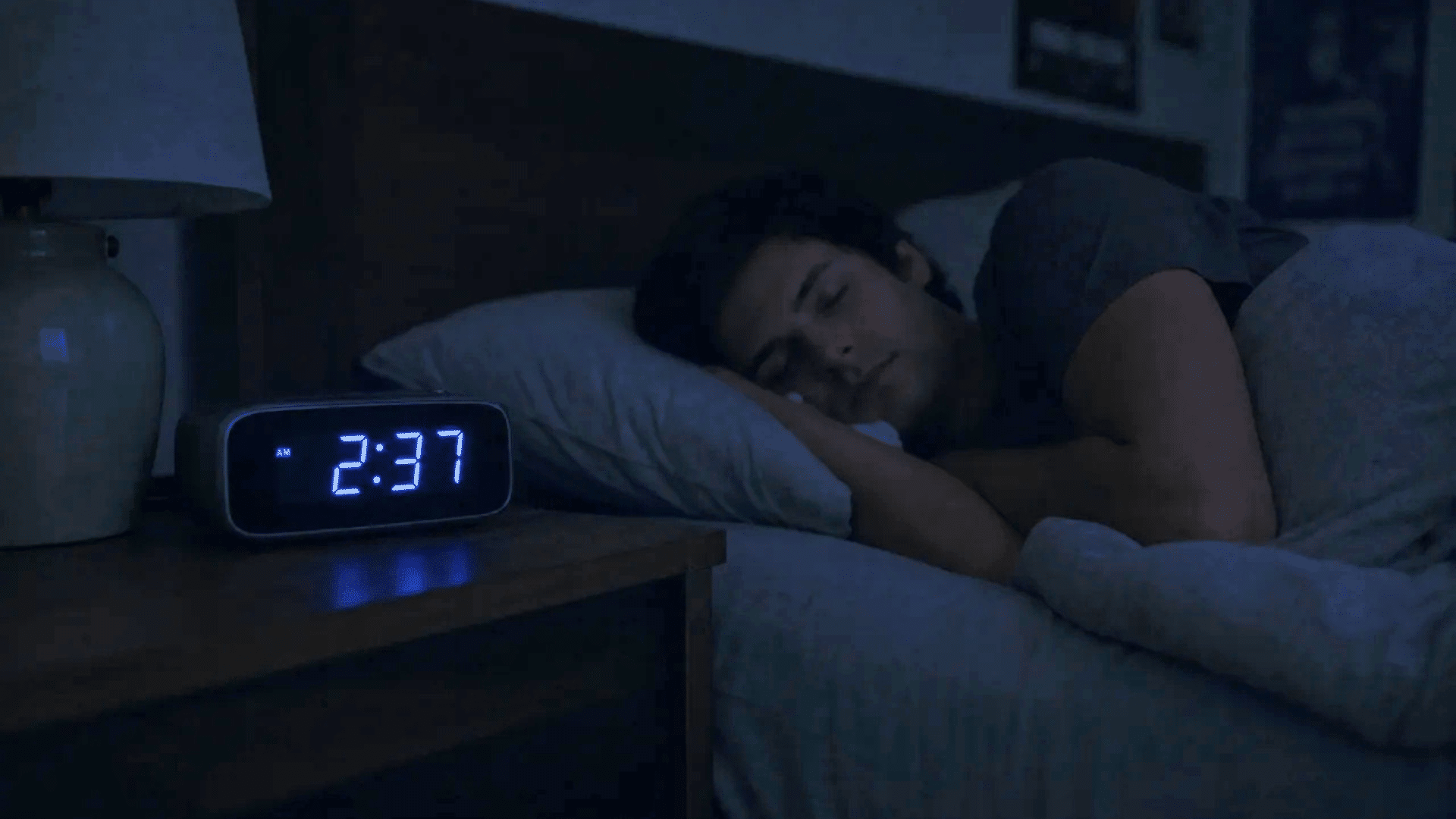 A person sleeping peacefully in a dark room with a glowing digital alarm clock on the nightstand.