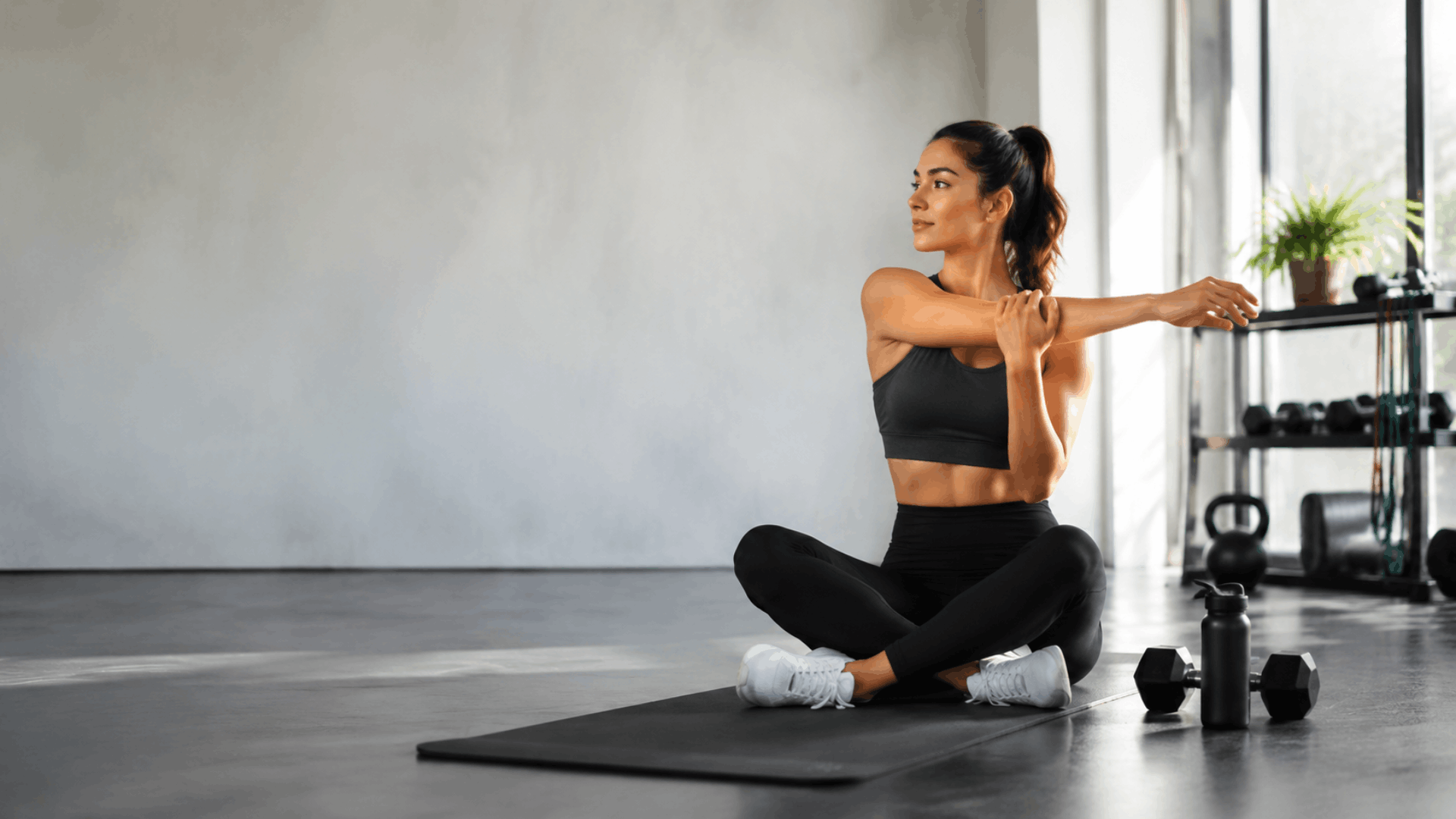 Woman stretching on yoga mat after workout in modern gym with dumbbells nearby