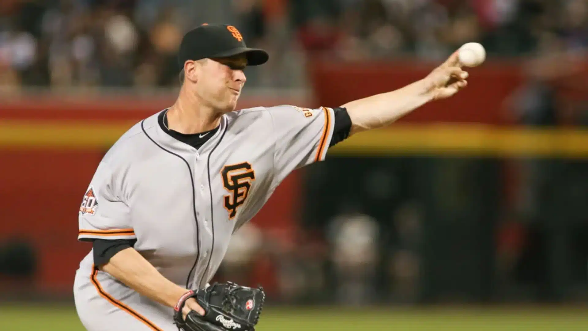 a baseball pitcher in a gray uniform throws a pitch during a game with his arm extended and the crowd blurred behind him