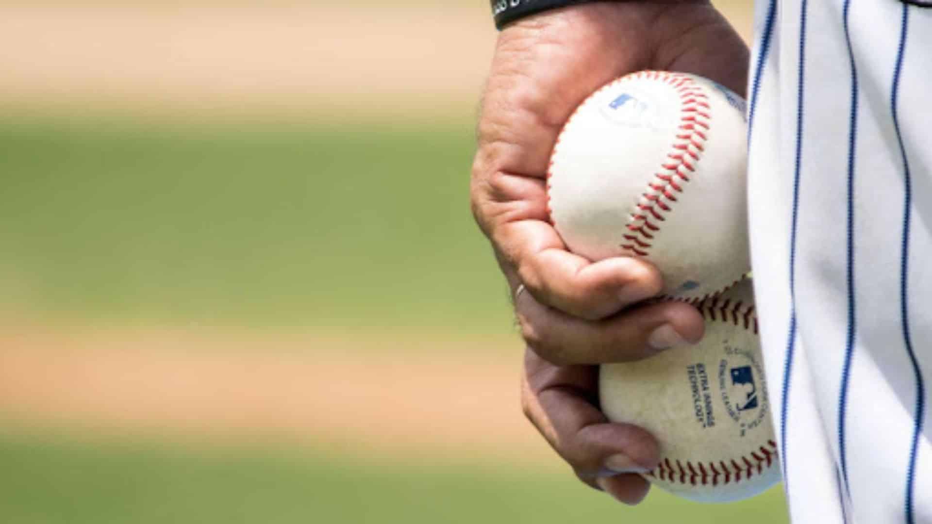 a close up of a baseball player’s hand gripping two baseballs at their side with a blurred green field in the background
