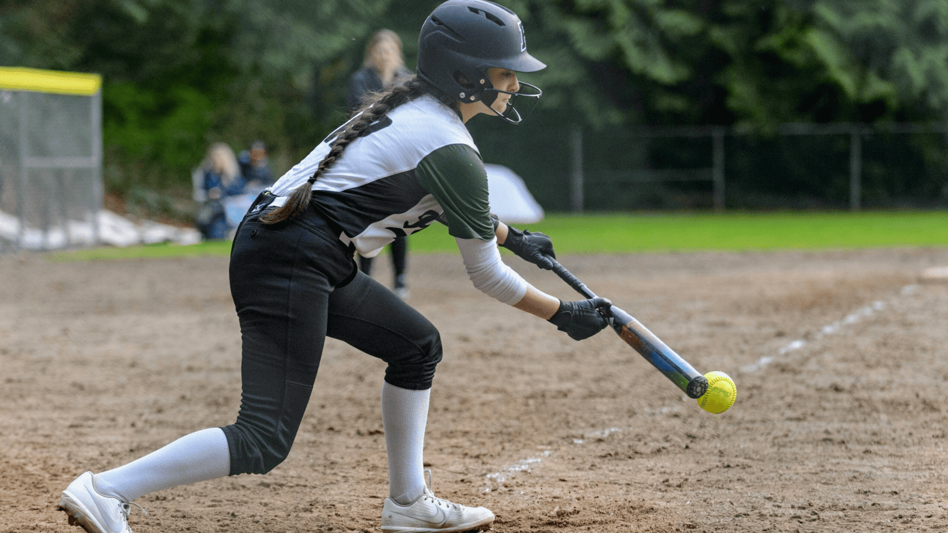 a softball player in a helmet bunting a yellow ball during a game on a dirt field with trees in background