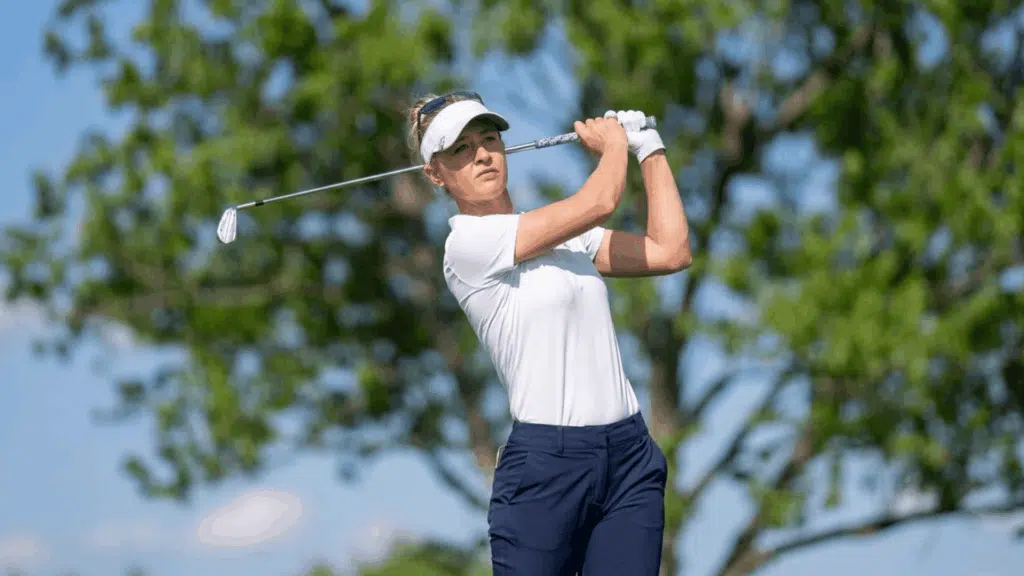 a woman in a white shirt and visor swinging a golf club on a sunny day with trees in the background