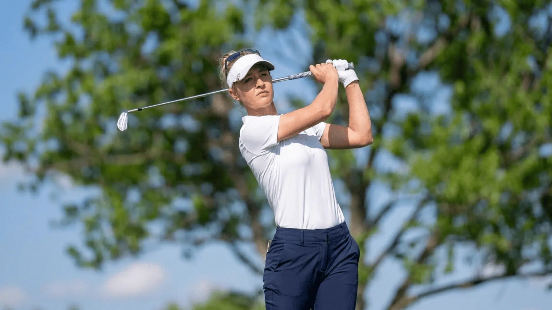 a woman in a white shirt and visor swinging a golf club on a sunny day with trees in the background
