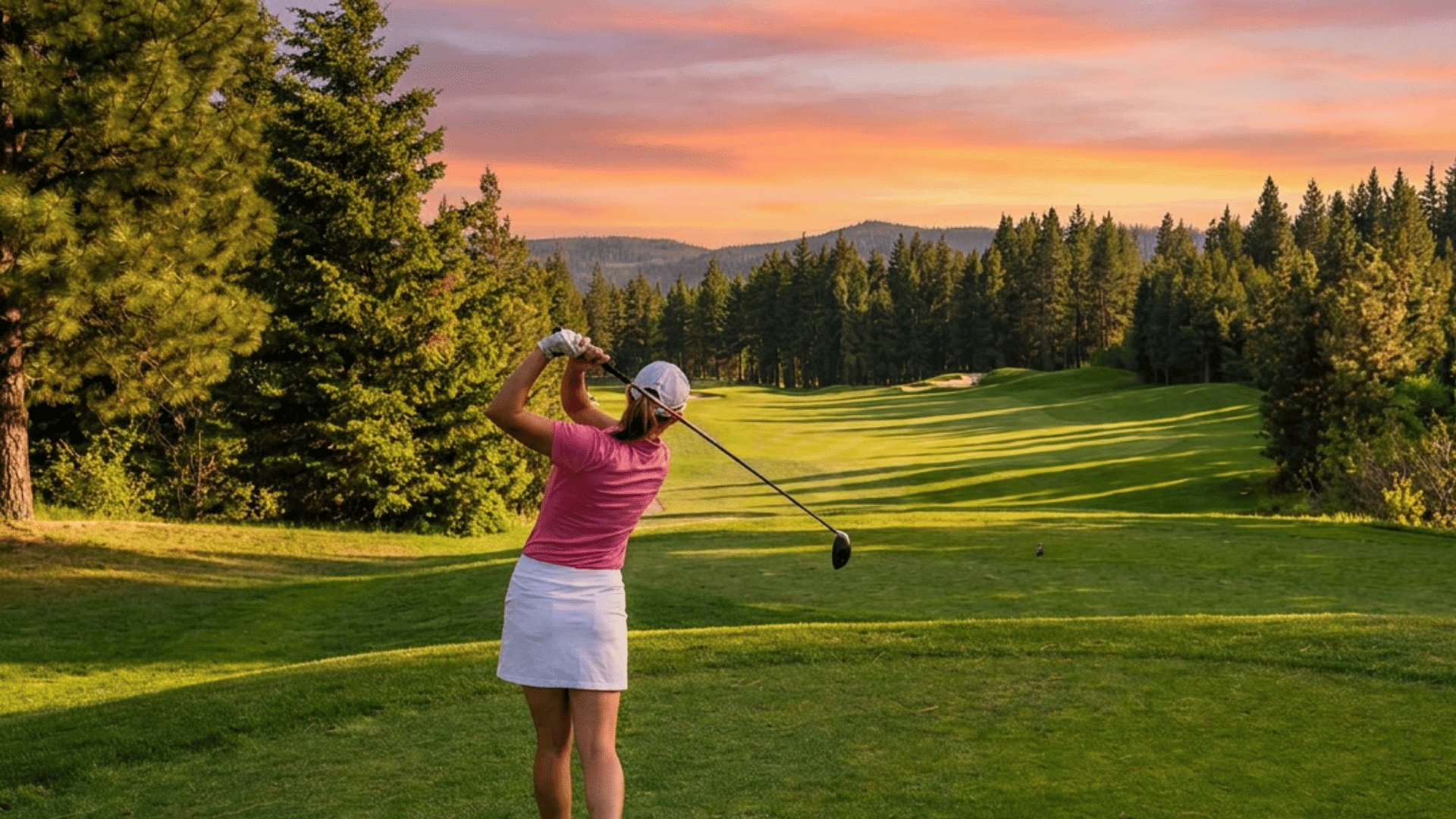 a woman swinging a golf club on a tree lined golf course with rolling hills during a colorful sunset sky