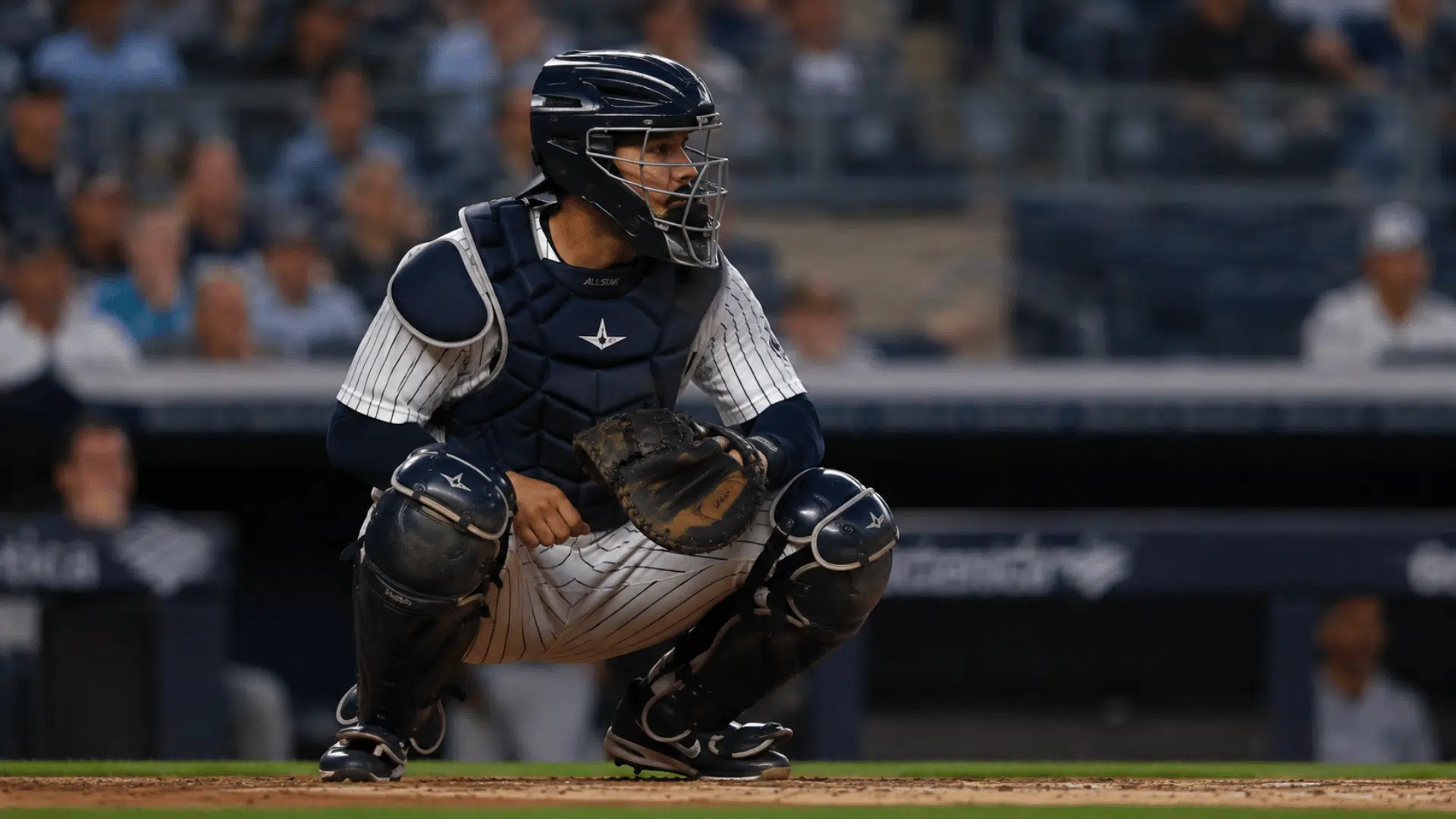 baseball catcher wearing full protective gear crouching on field during game with stadium crowd behind him