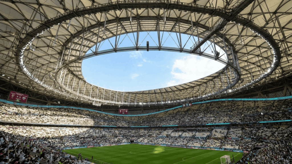 crowded football stadium beneath a curved roof with sunlight shining over the green field and fans
