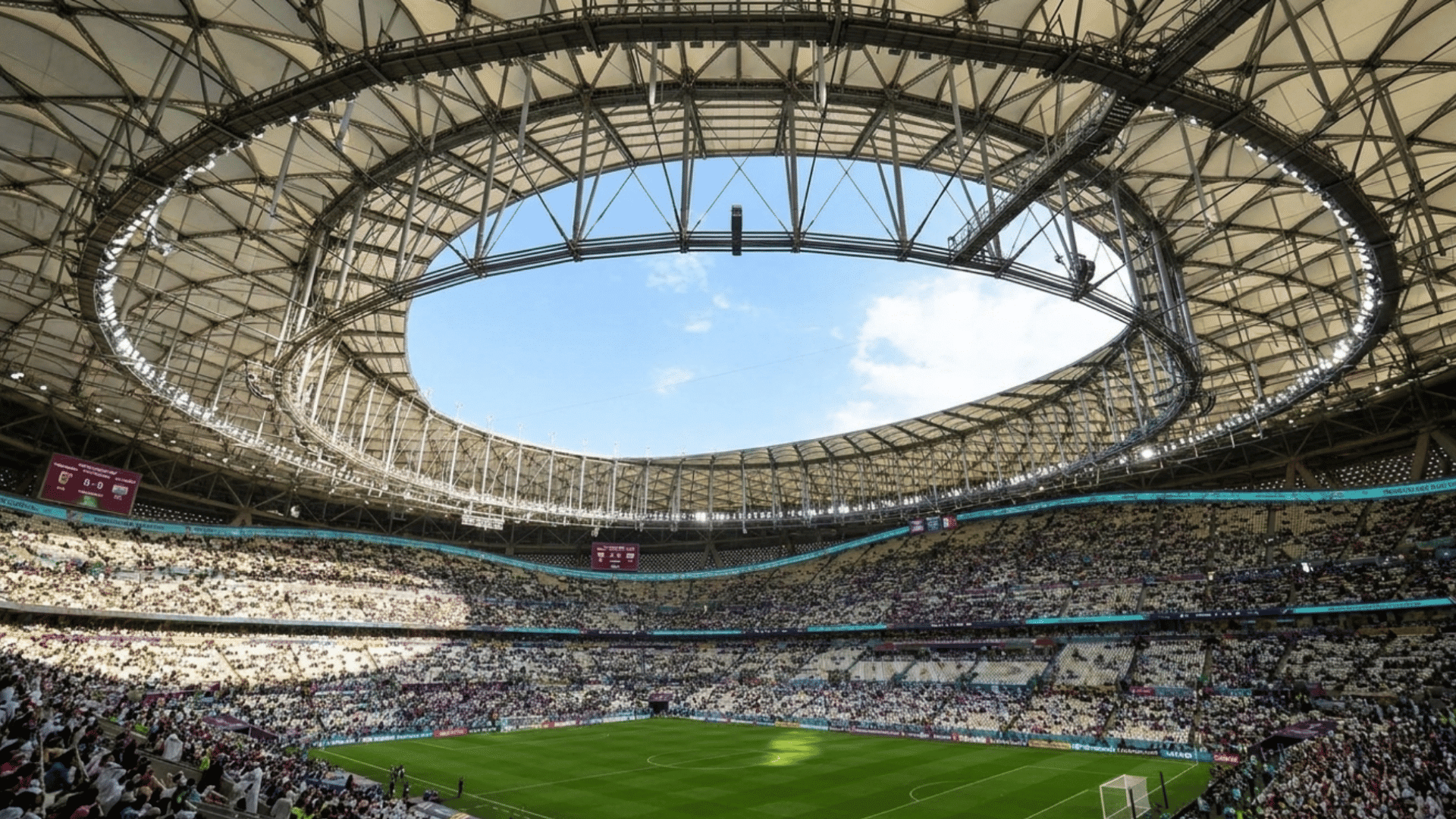 crowded football stadium beneath a curved roof with sunlight shining over the green field and fans