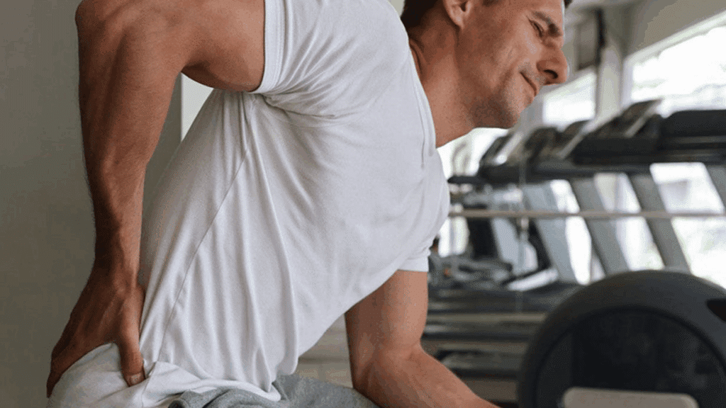 man holding lower back in pain after workout while sitting inside a gym during recovery time