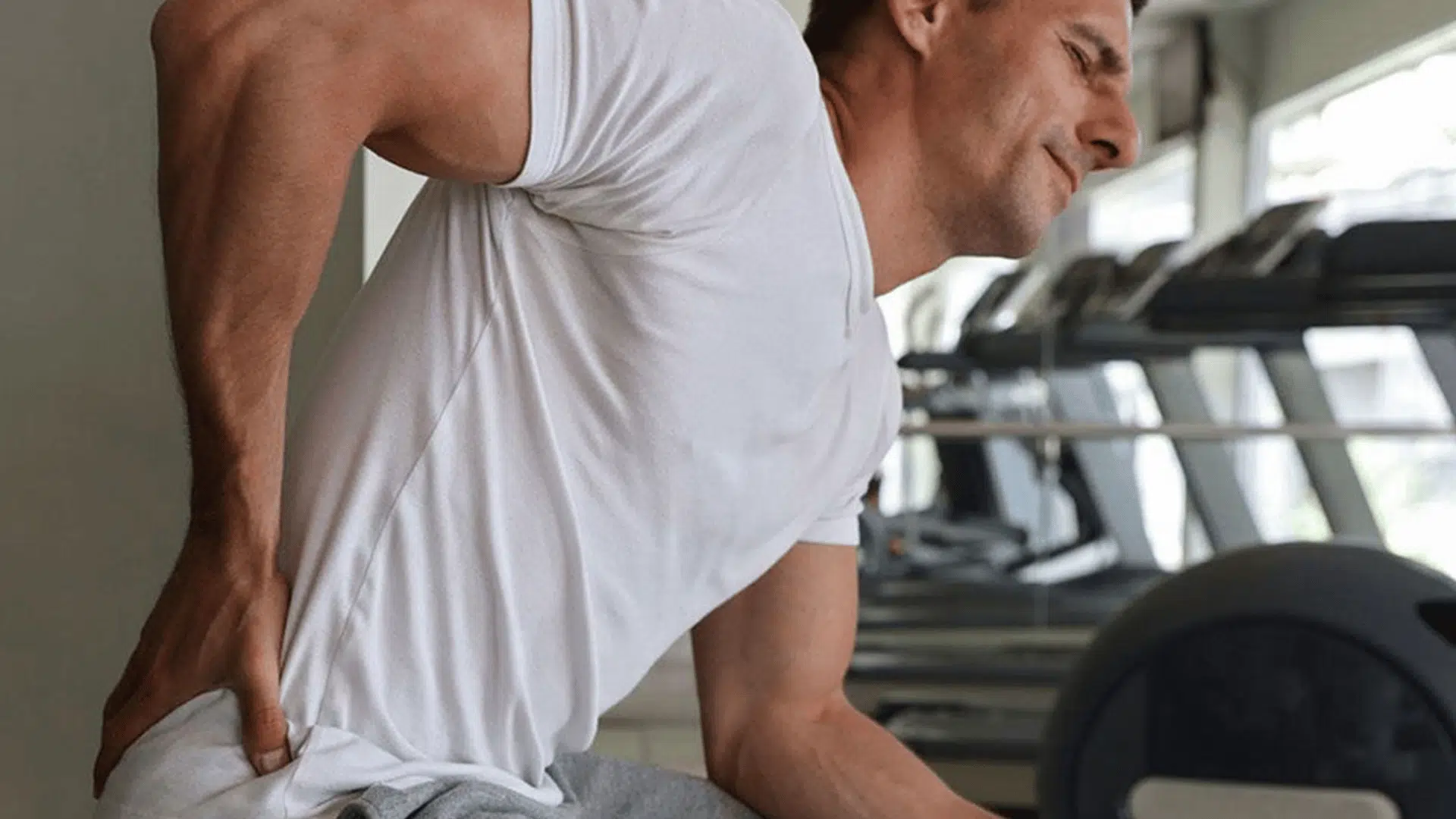 man holding lower back in pain after workout while sitting inside a gym during recovery time