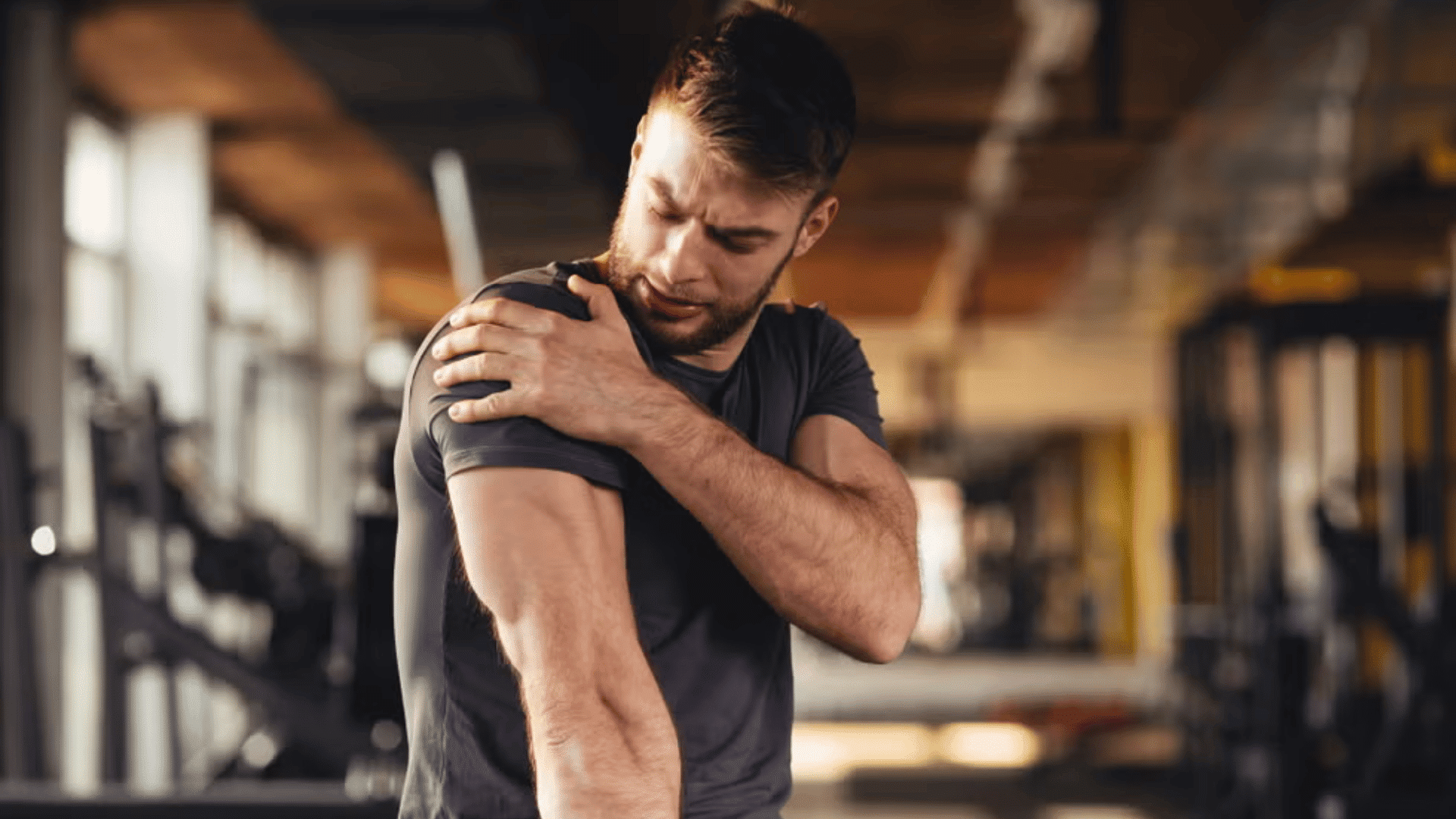 man holding sore shoulder after workout while sitting inside a gym and checking muscle pain