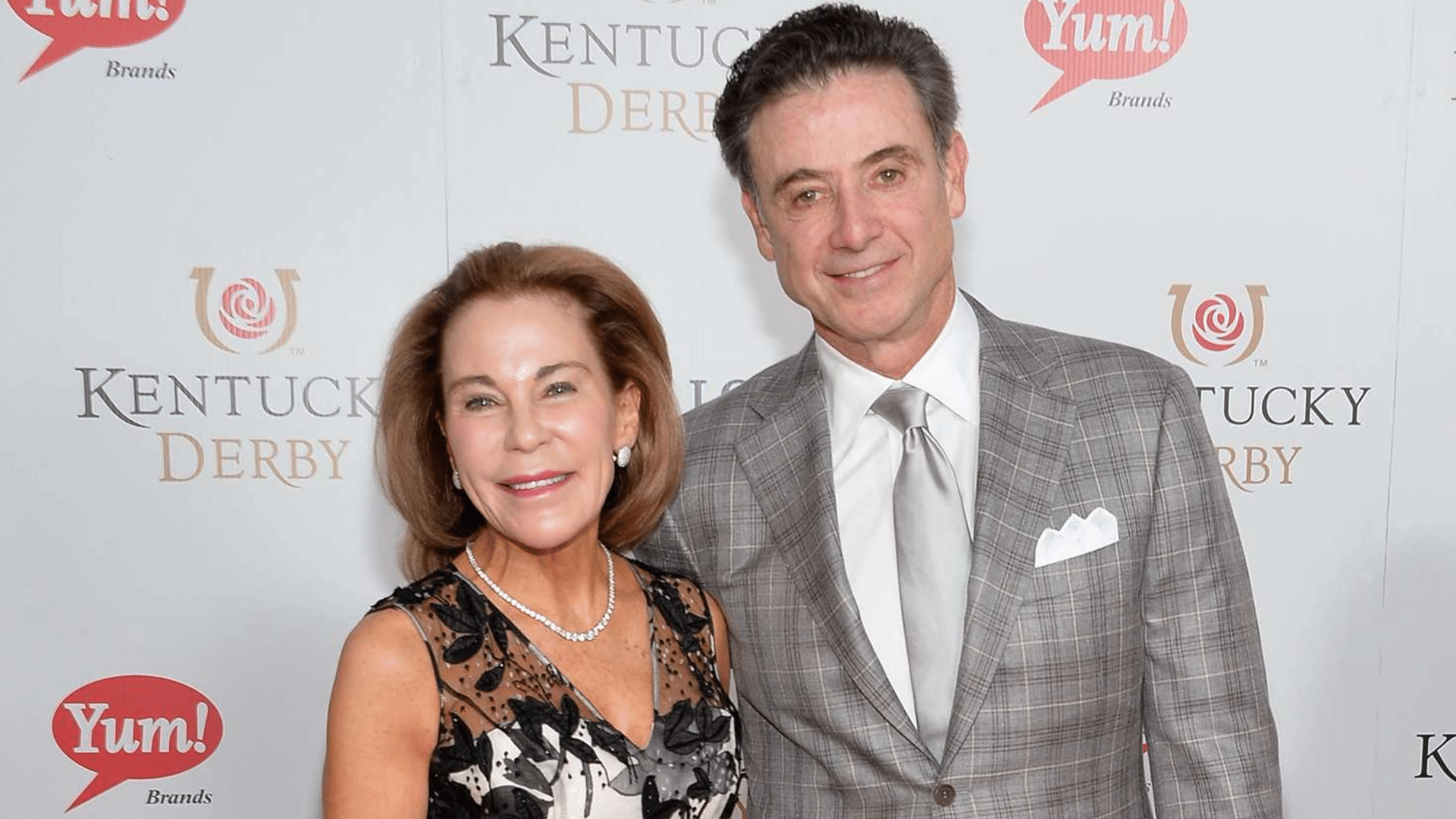 man in gray suit standing beside woman in floral dress posing at kentucky derby event backdrop