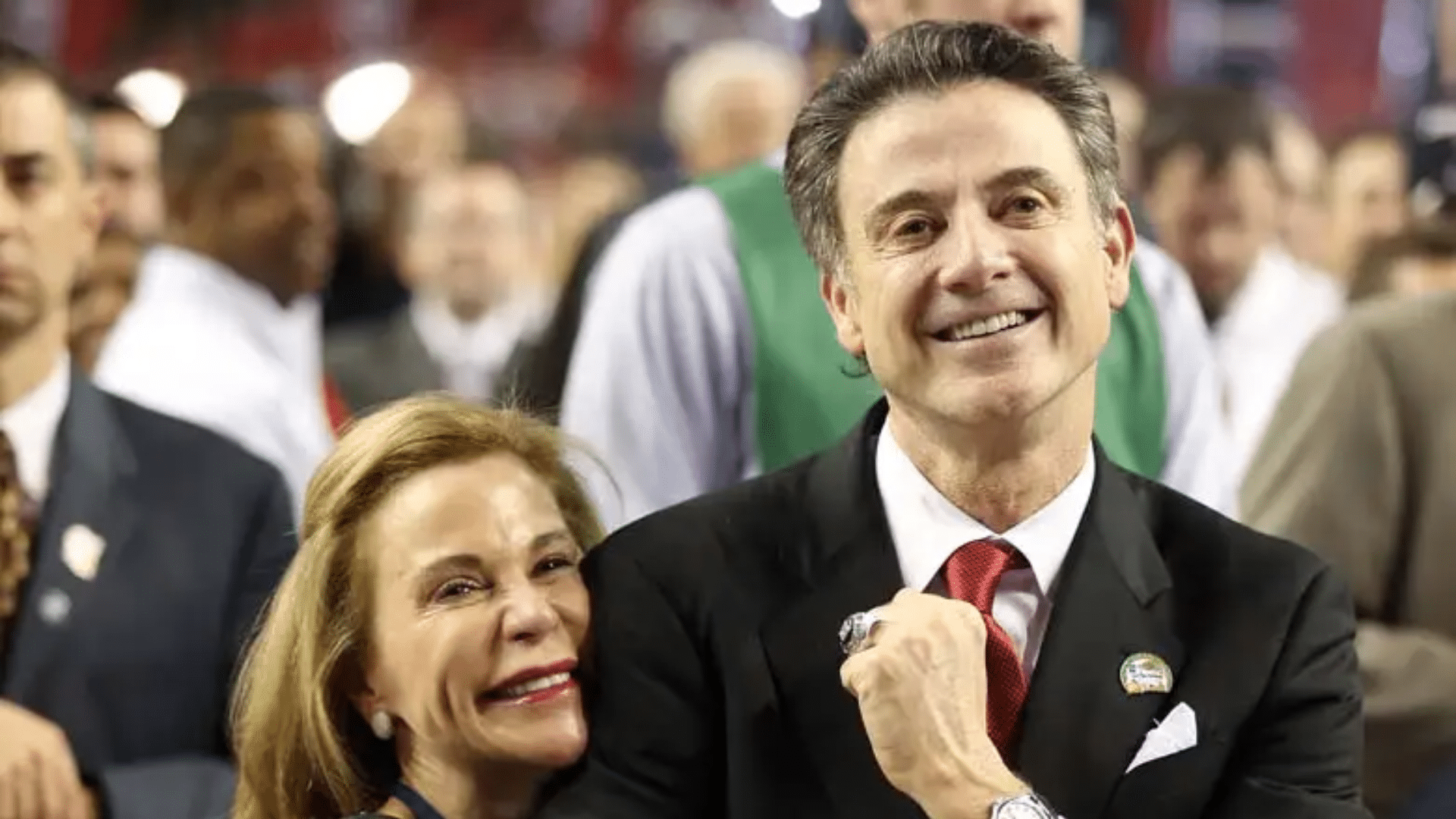 smiling man in black suit standing beside woman hugging him in crowded sports arena setting