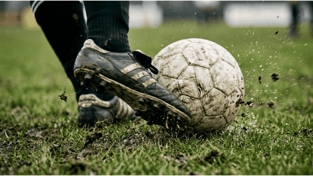 soccer player kicking a muddy ball on wet grass during an outdoor match with dirt flying through the air
