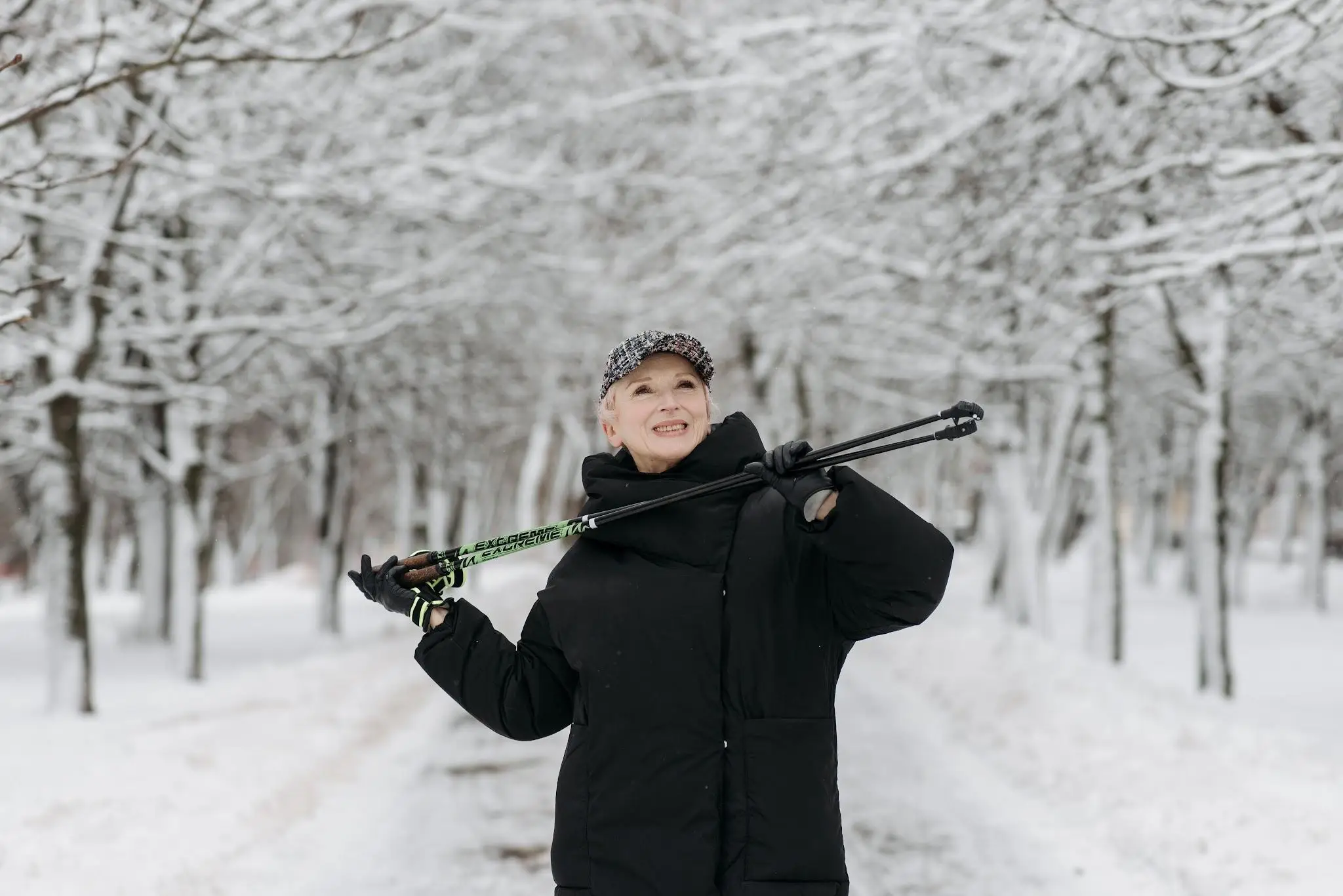Person in black winter coat holding trekking poles in snowy tree-lined path