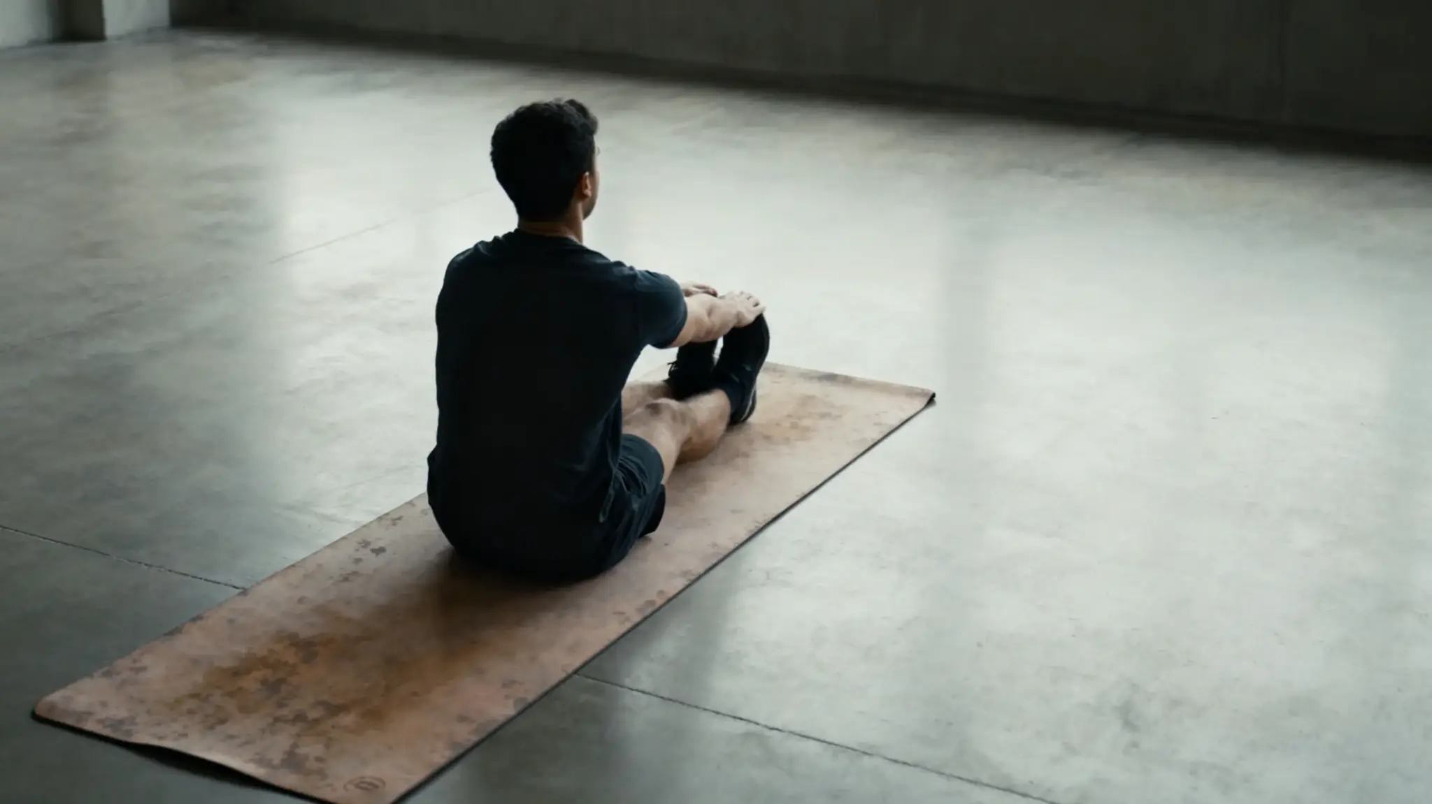 Man stretching on brown yoga mat in spacious indoor studio with concrete floor