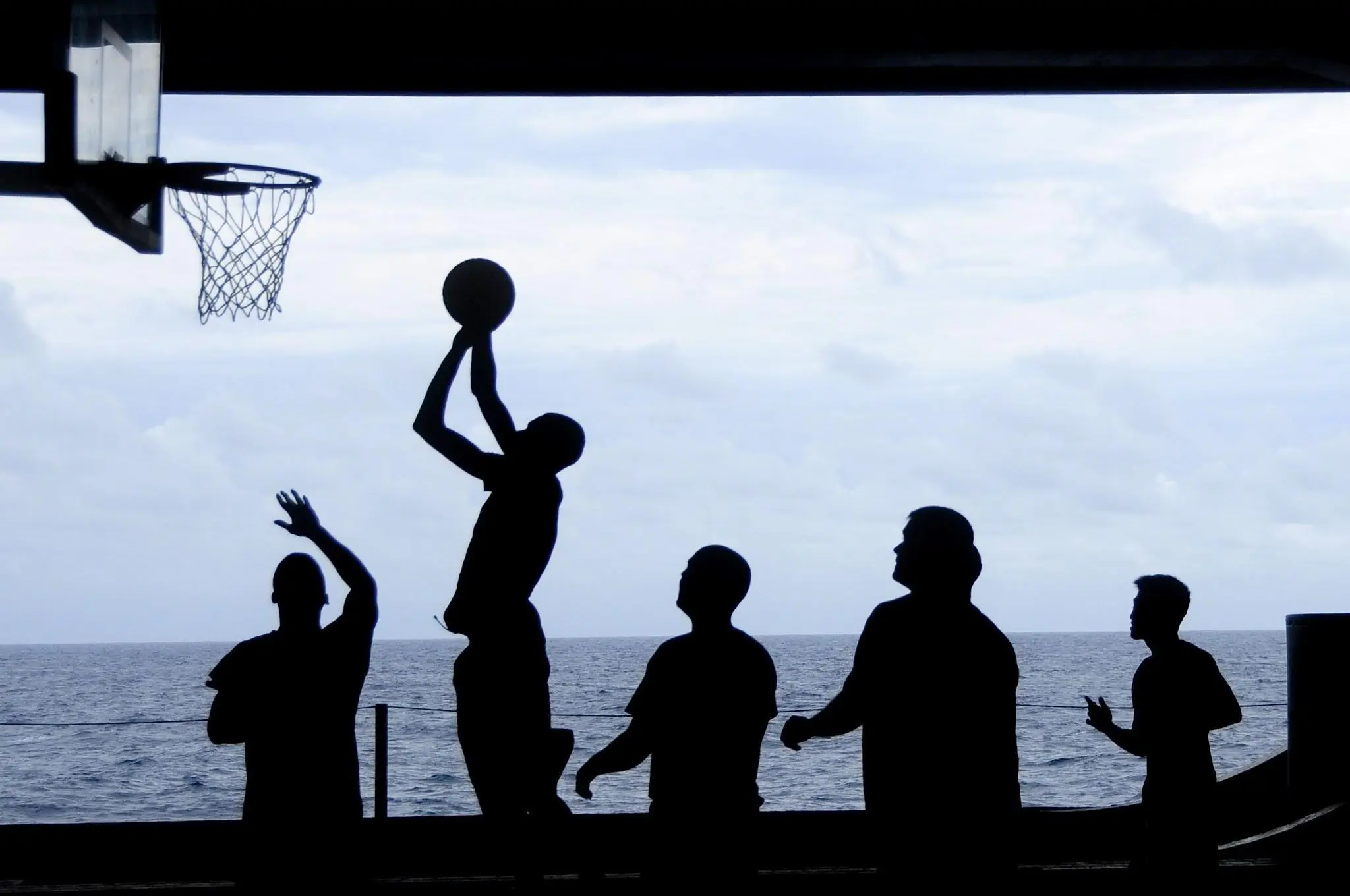 Silhouetted people playing basketball on an outdoor court near the ocean.