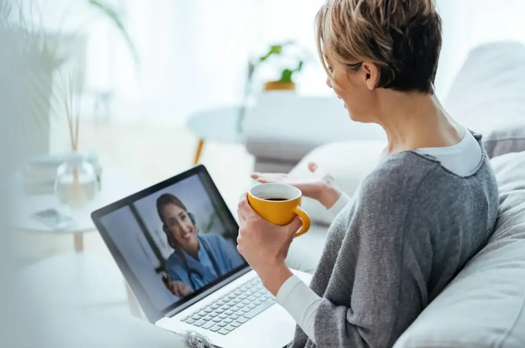 Woman having video call with doctor on laptop while holding yellow coffee mug indoors