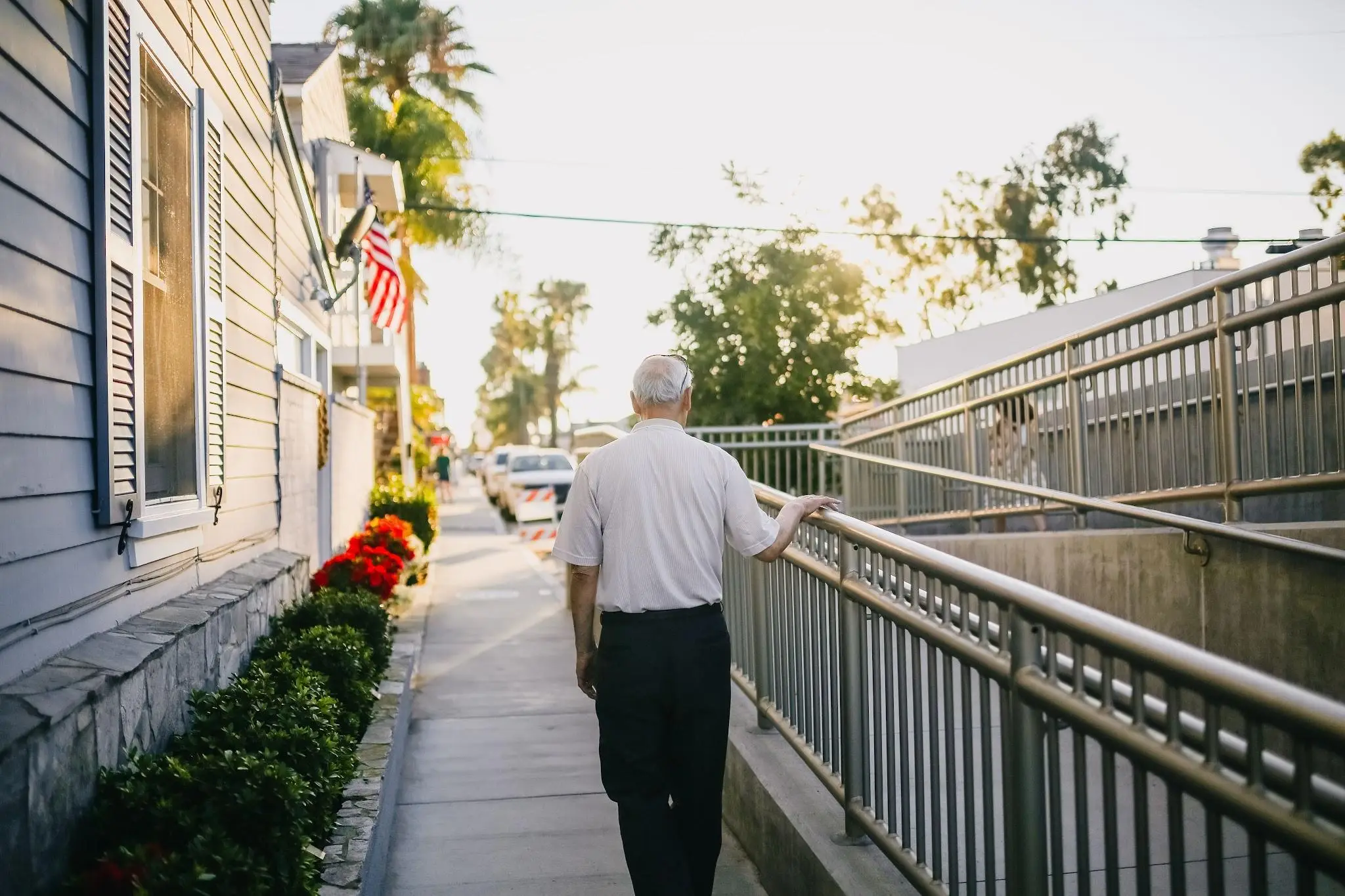 Elderly man walking along sidewalk with hand on railing in suburban neighborhood during sunset